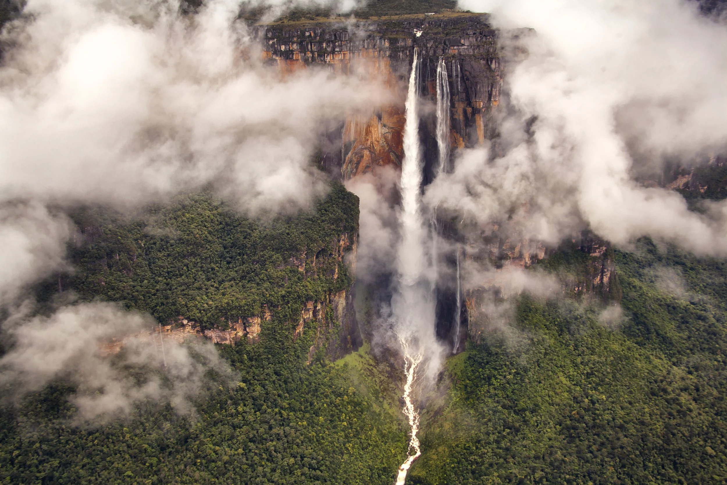 Aerial view of a tall waterfall cascading down a cliff surrounded by dense green forest, with mist and clouds partially obscuring the scene.