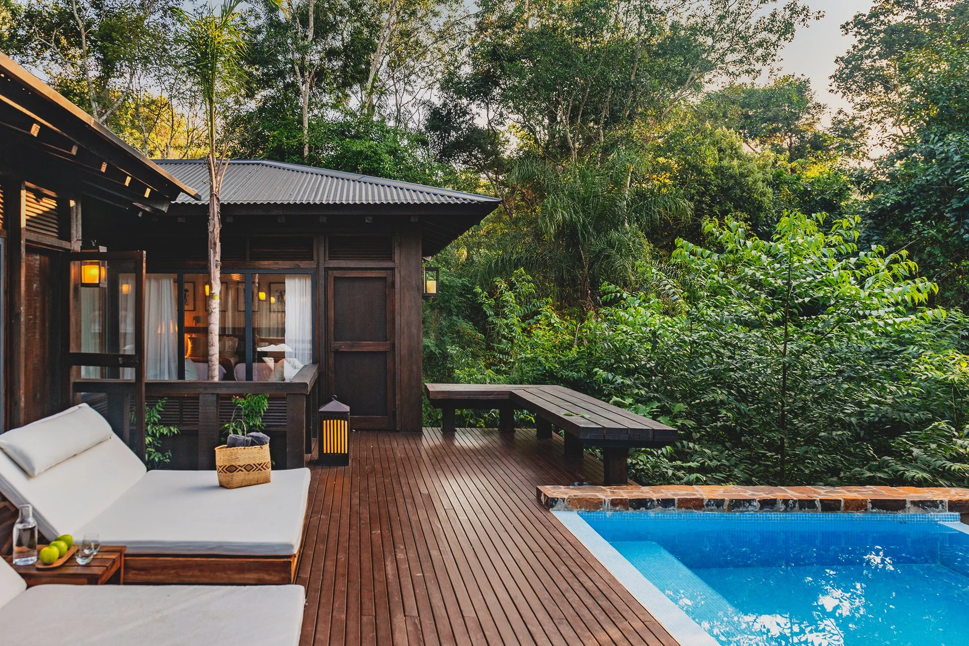 Wooden deck with outdoor furniture and pool, surrounded by lush green trees and a cabin with interior lights visible through glass doors, Iguazu Falls, Argentina