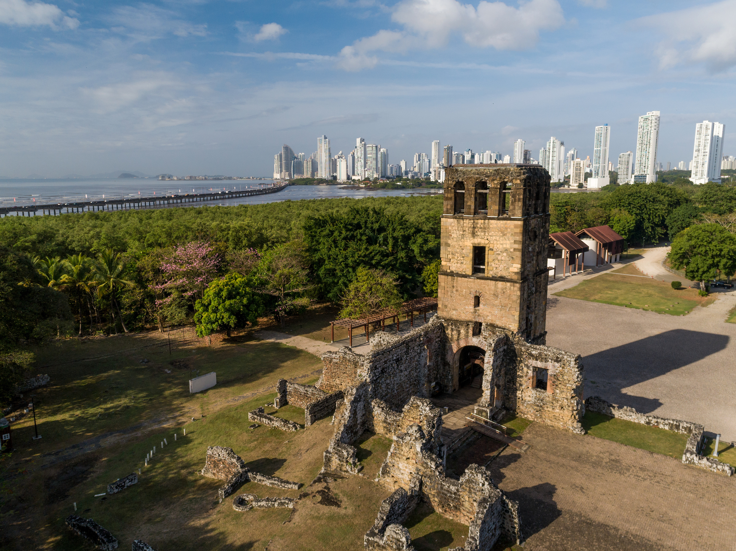 Aerial view of a historic stone tower with ruins in the foreground, surrounded by trees, with a modern city skyline in the background, near a body of water under a partly cloudy sky.