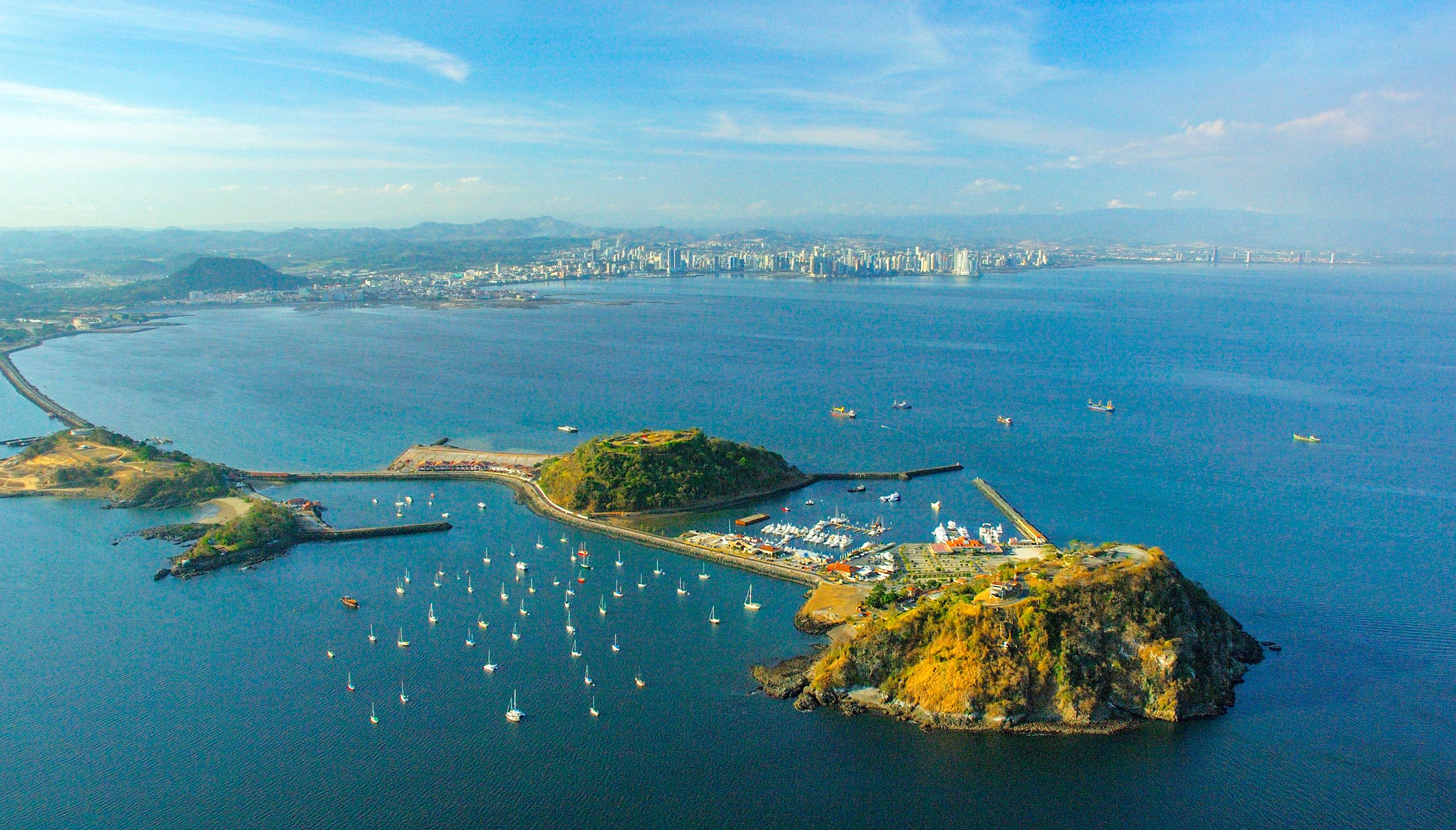 Aerial view of Gunkanjima, a small island with a harbor, boats, and surrounding water, with a cityscape and mountains in the background.