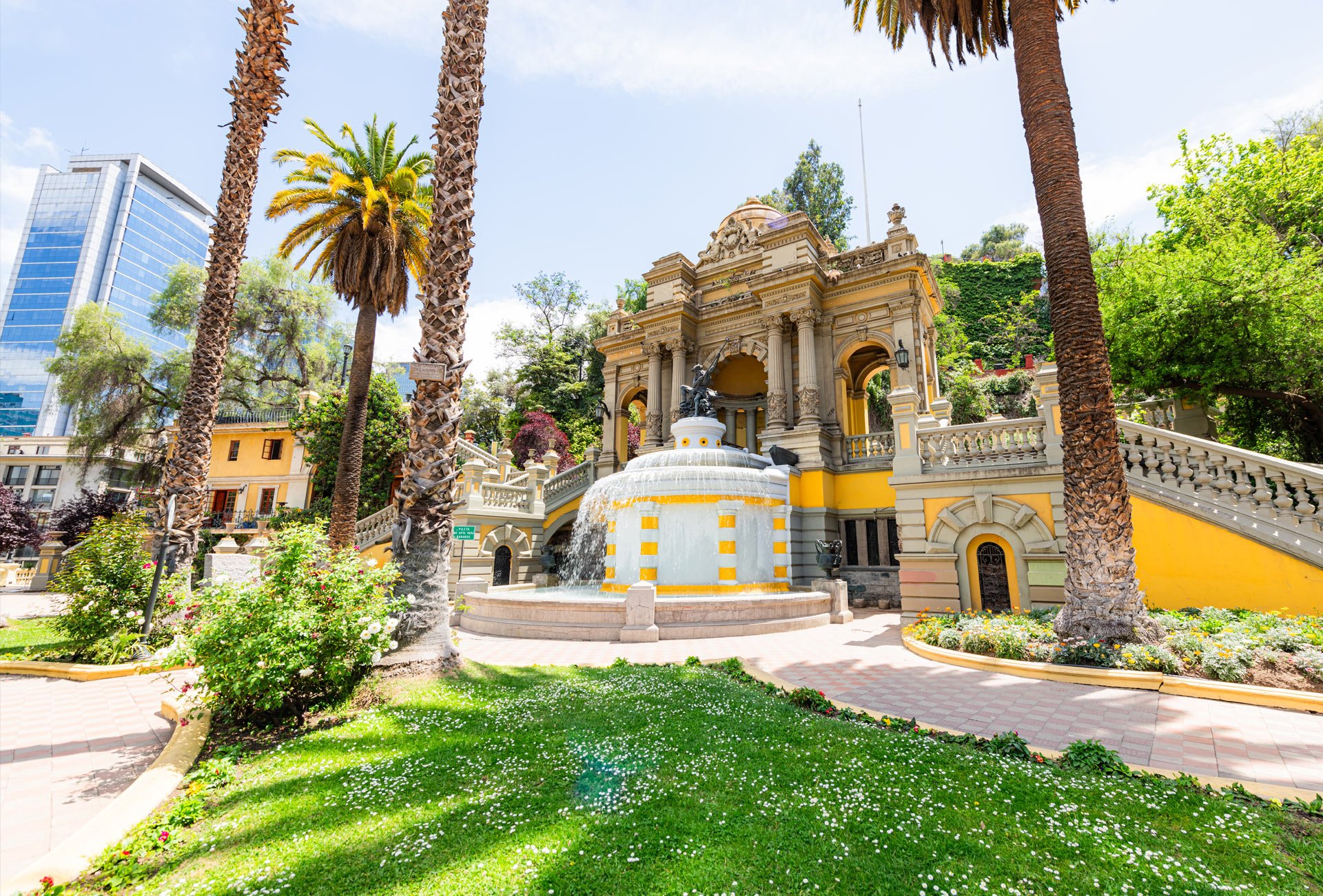 Historic fountain on a sunny day in a lush garden with tall palm trees, surrounded by colorful flowers and green grass, with a modern glass building in the background. Santiago de Chile, Chile.