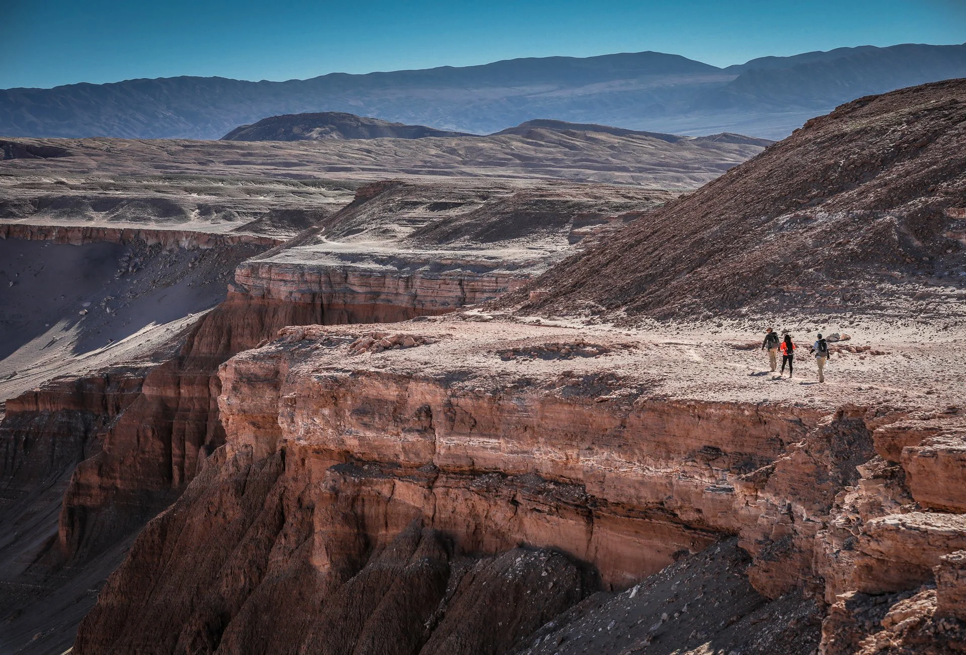 Four hikers walking along the edge of a canyon with layered rock formations and distant mountains under a clear blue sky. Peru.
