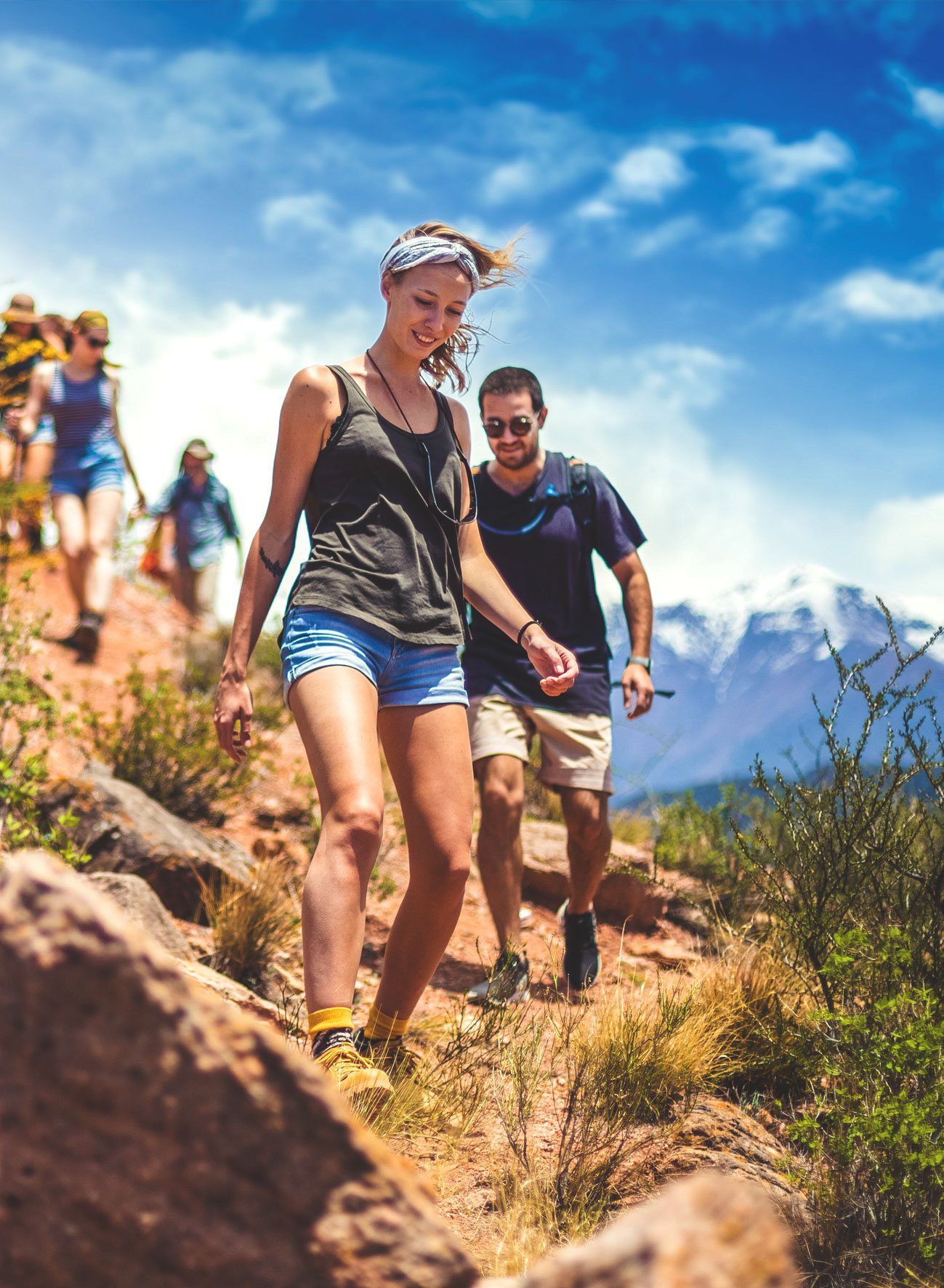 Group of young hikers walking on a rocky trail in a mountainous area with snow-capped peaks in Argentina with on a sunny day and blue sky.