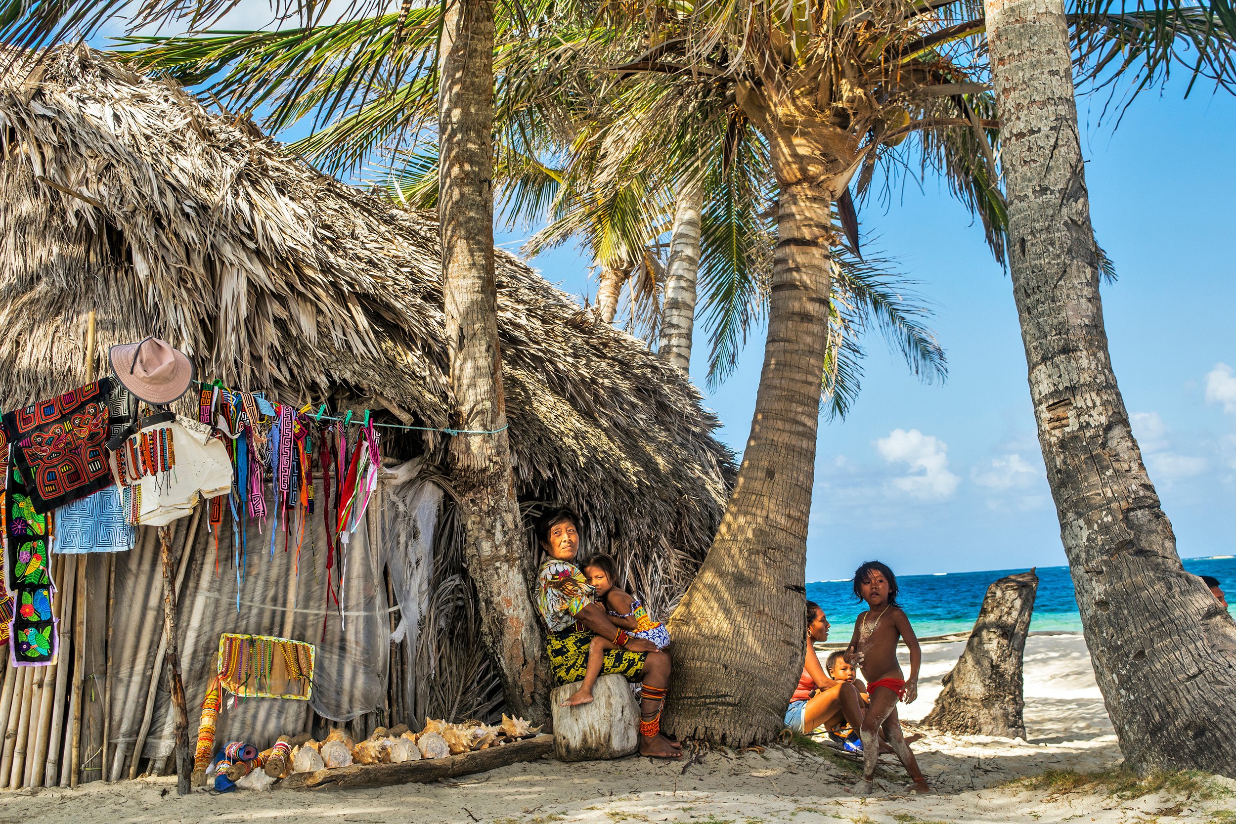 Indigenous people gathered near a thatched hut with colorful handicrafts hanging, on a beach with palm trees and the ocean in the background.