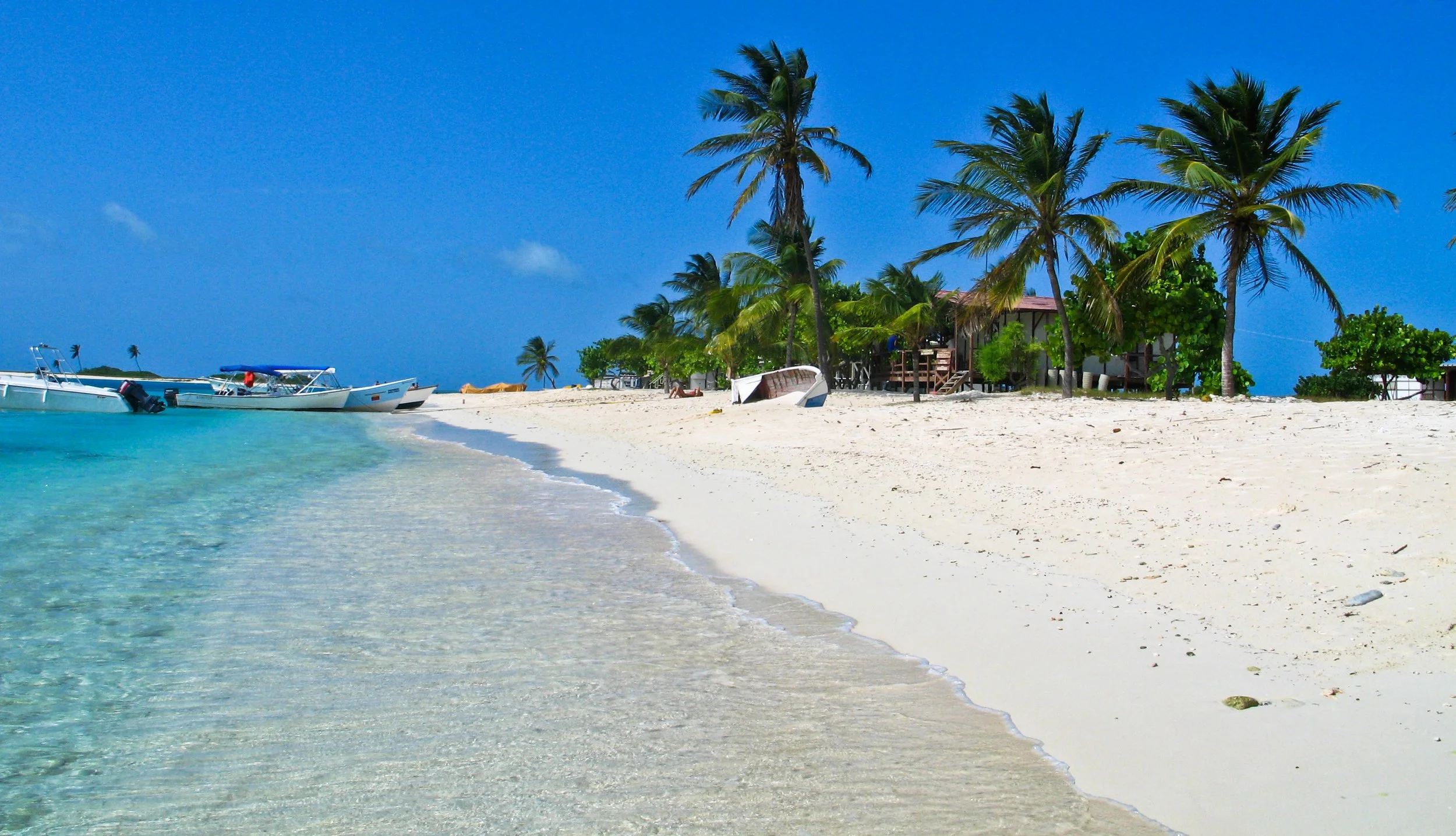 Tropical beach with turquoise water, white sand, palm trees, and boats anchored near the shore under a clear blue sky.