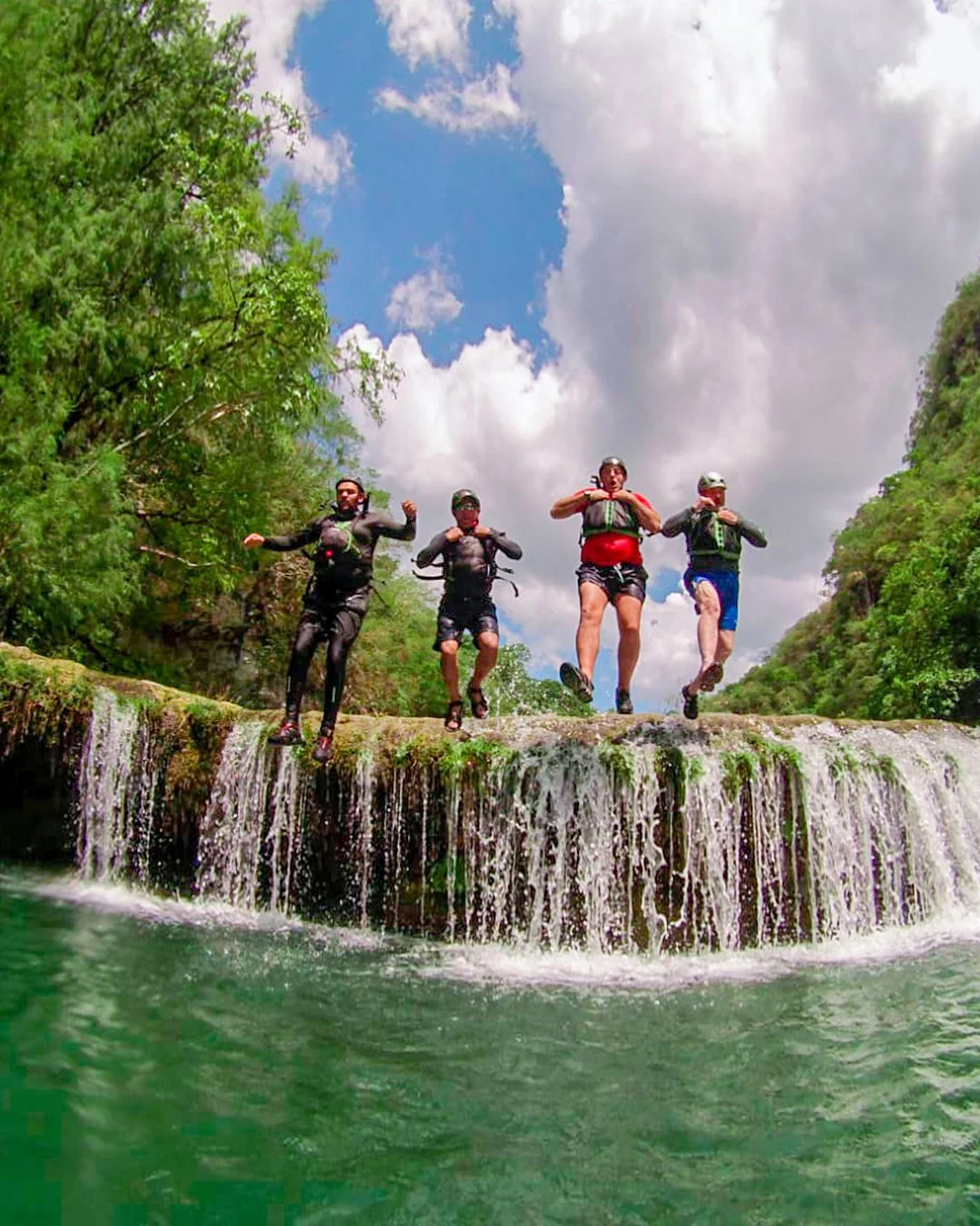 Four people, wearing life jackets and water gear, jumping off a small waterfall into a river surrounded by green trees and blue sky with white clouds.