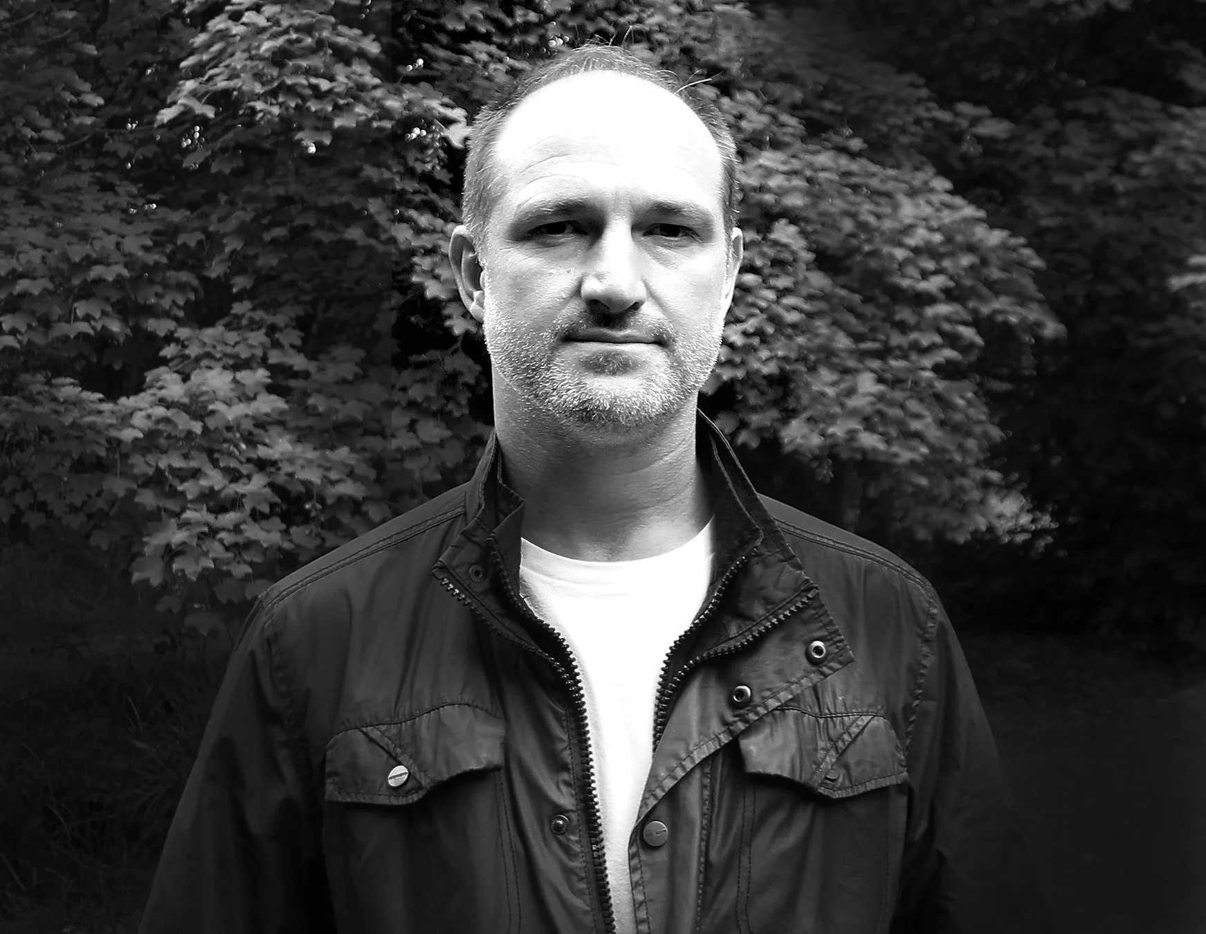 A black and white photo of a middle-aged man with short hair and a beard, standing outdoors in front of dense trees. He is wearing a dark jacket over a light shirt.