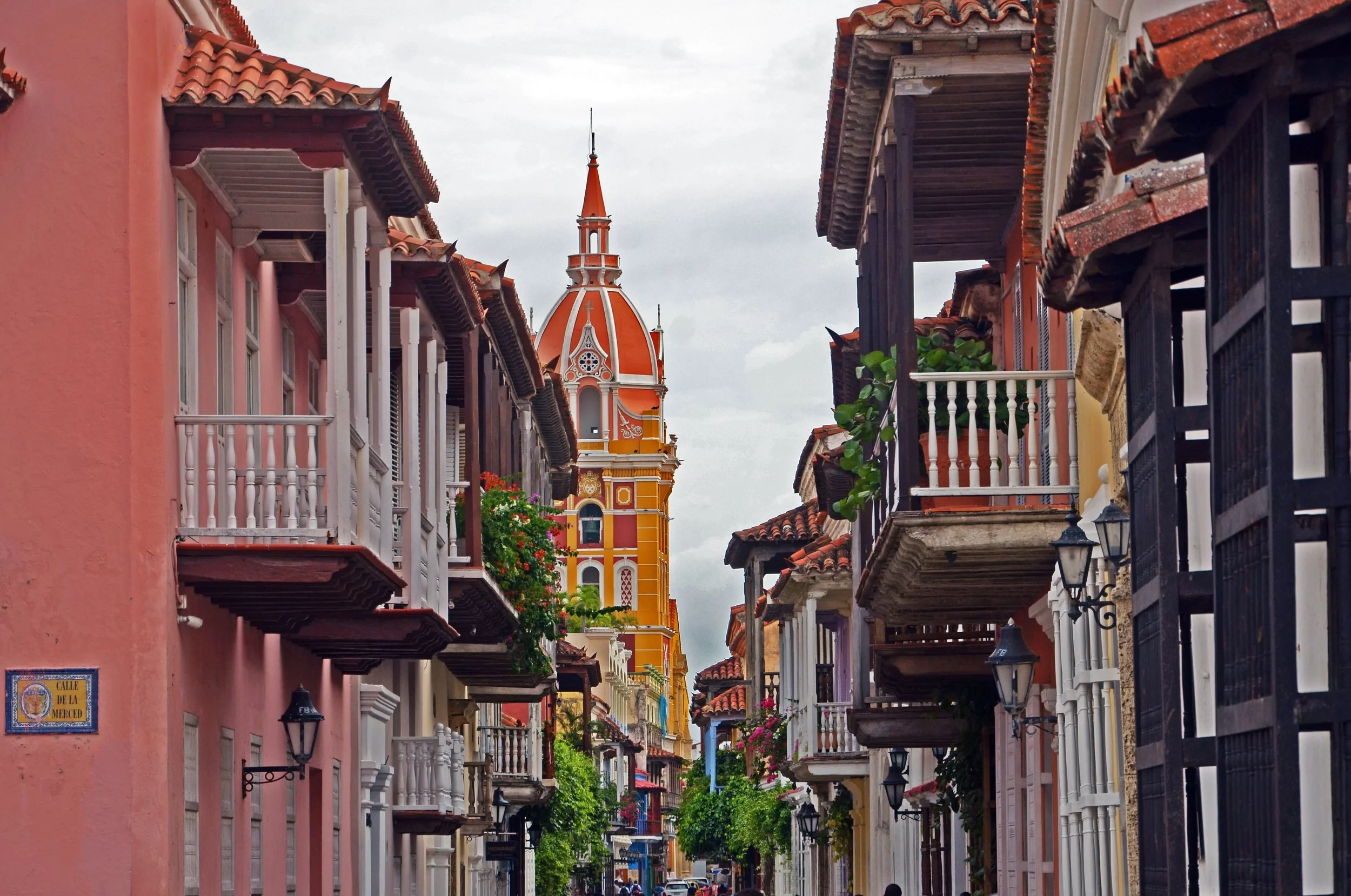 Colorful street with colonial-style buildings and balconies, and a tall orange church with a pointed spire in the background.