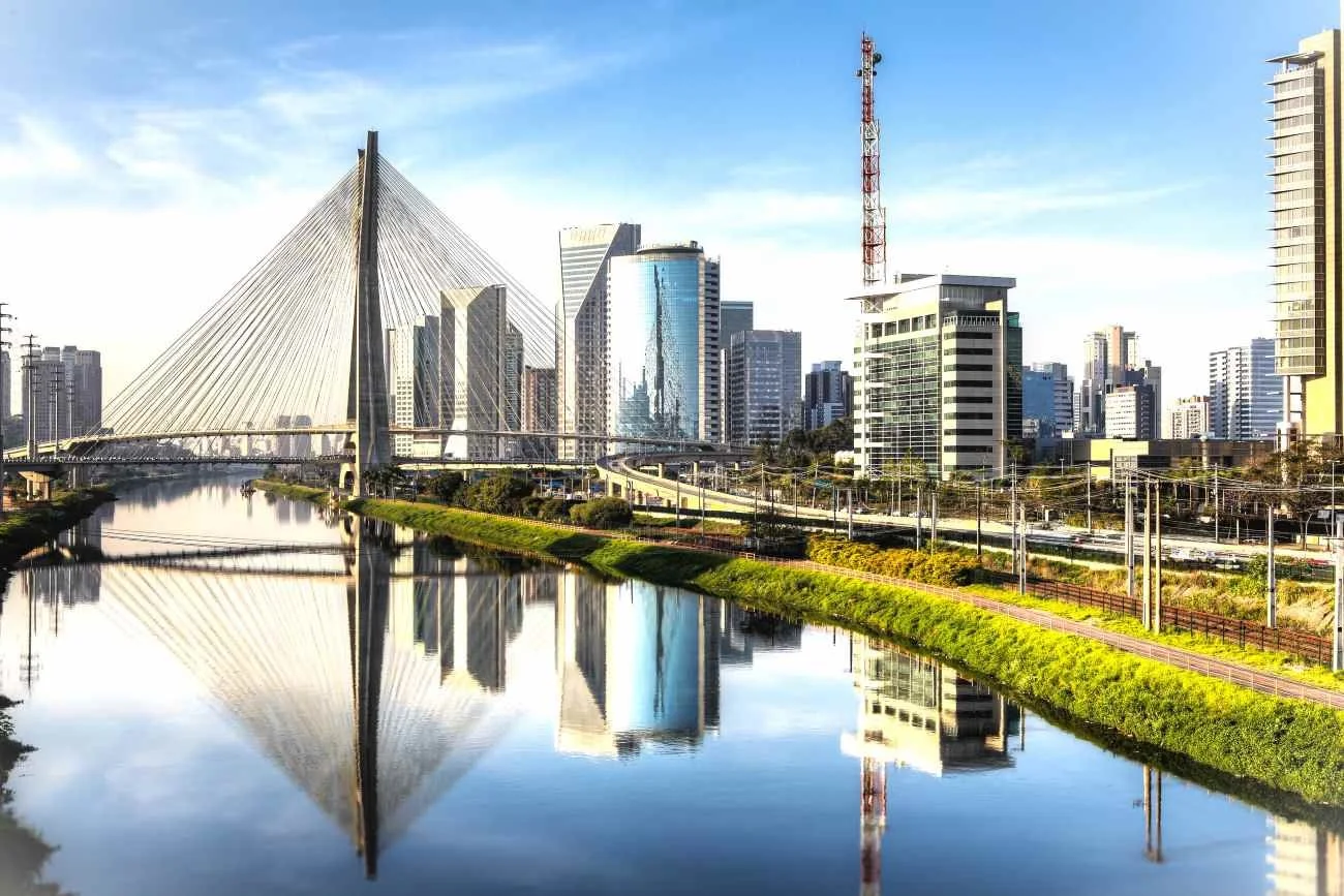 City skyline with modern high-rise buildings, a cable-stayed bridge, and a river reflecting the skyline under a blue sky.