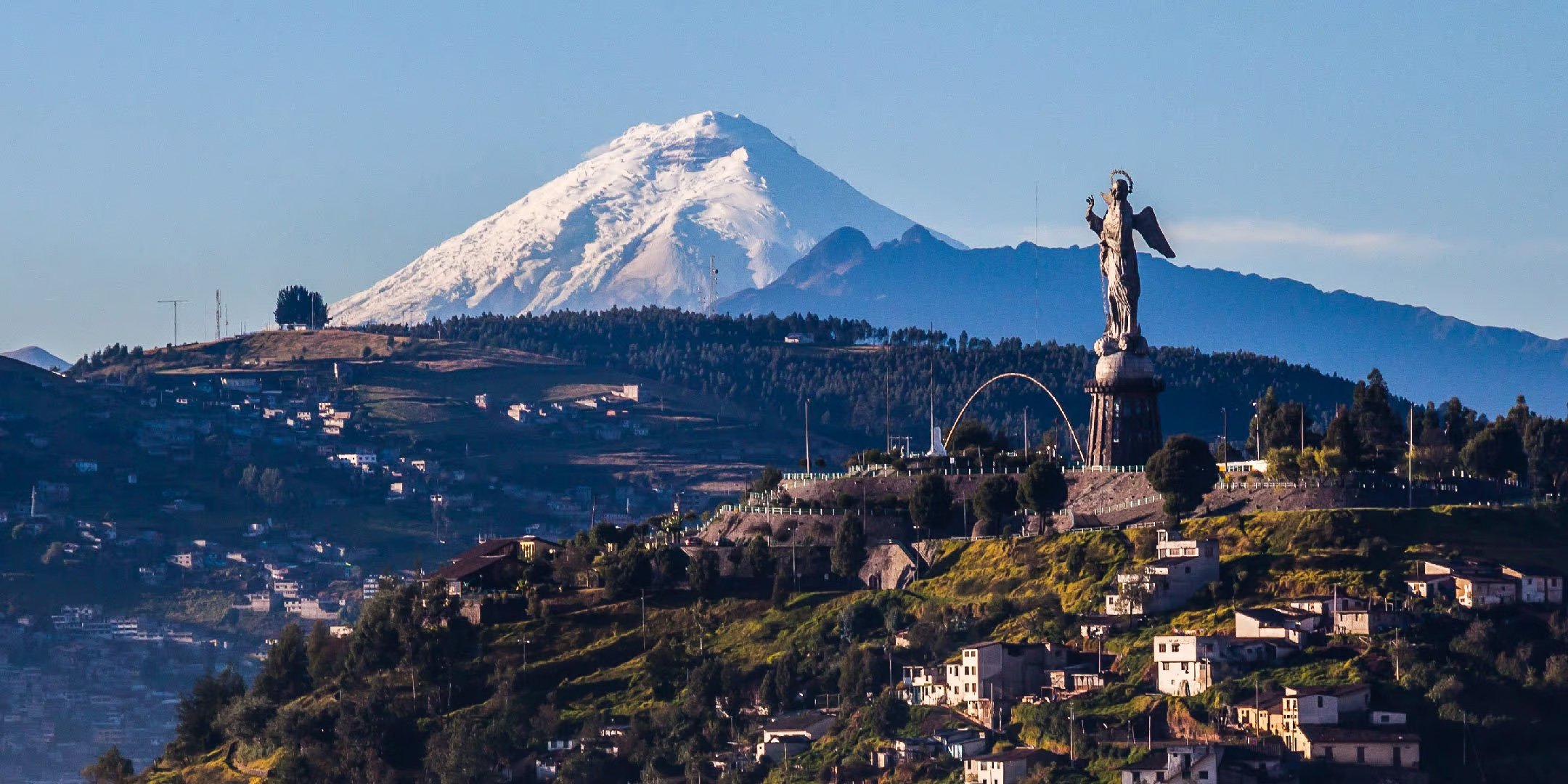 A landscape view of a hillside city with a large statue and mountain in the background, snow-capped mountain peak visible. Ecuador.