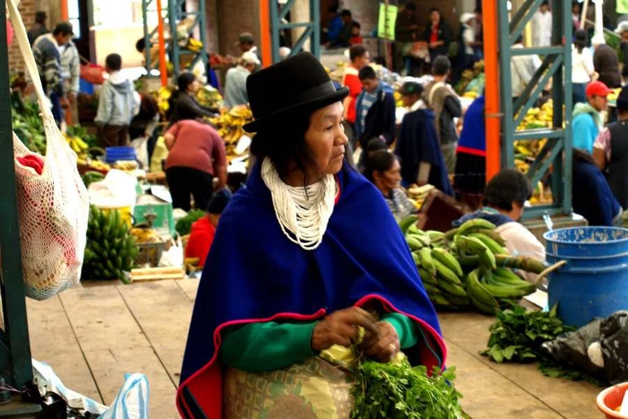 A woman in traditional clothing and a hat at a bustling outdoor market with numerous people, fruits, and vegetables in the background.