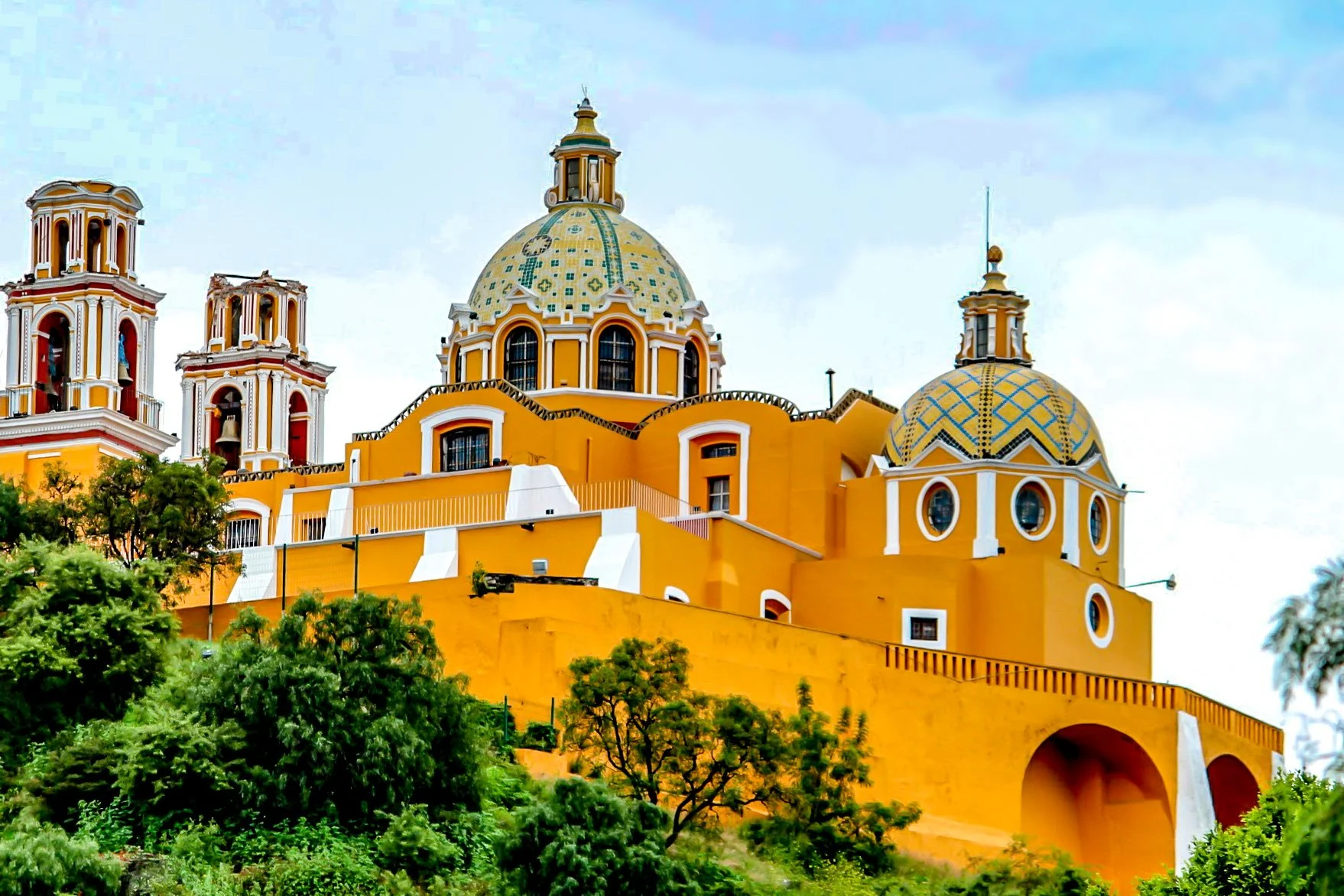 Colorful yellow church with domes and bells, surrounded by green trees, under a cloudy sky.