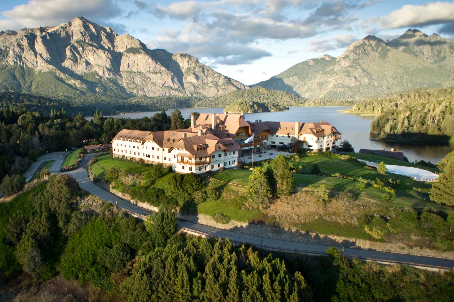 A large, multi-story building with brown roofs is situated on a hill overlooking a lake with mountains in the background. The scene is surrounded by green trees and a winding road. The sky is partly cloudy. Argentina.