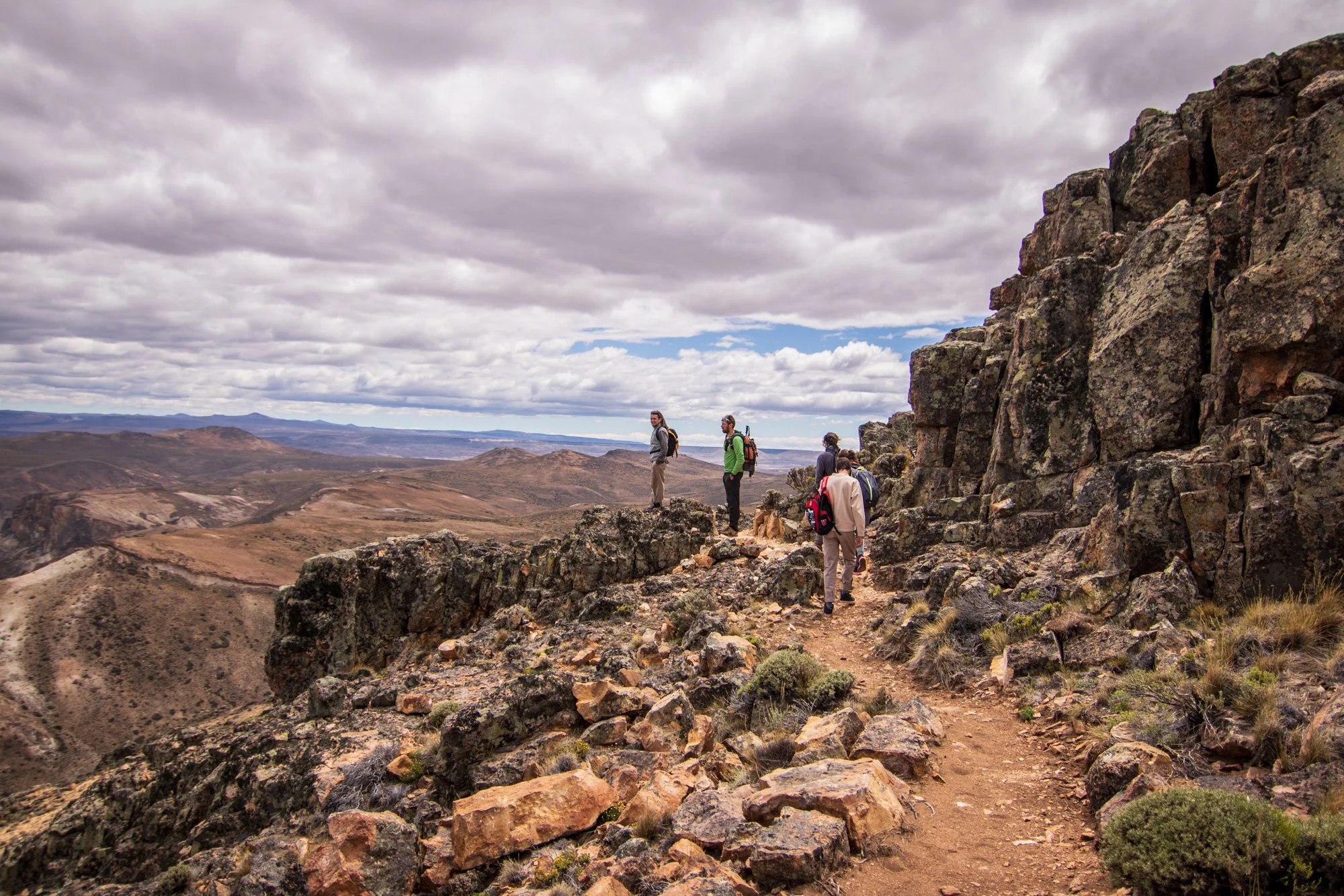 Group of hikers walking along a rocky trail near a cliff with a view of rolling hills and mountains in the distance, under a cloudy sky.