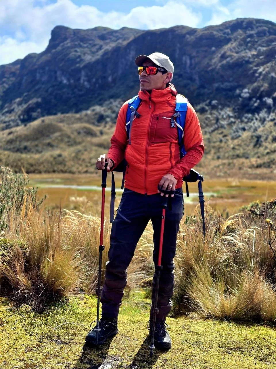 A man dressed in outdoor hiking gear, including a red jacket, black pants, and a cap, stands with trekking poles in a mountainous landscape with grassy terrain in the Andes mountain range in the background, Ecuador.