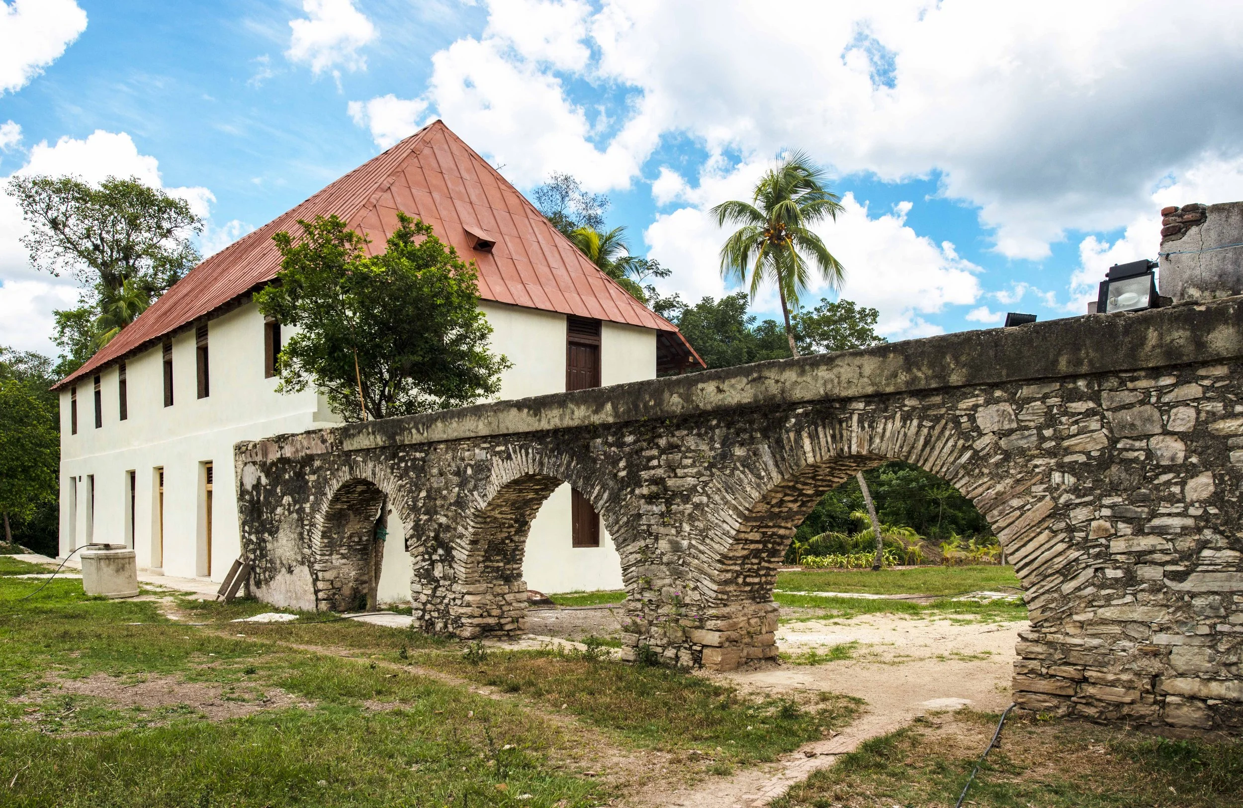 A historic stone aqueduct with three arches next to a white building with a red roof, surrounded by palm trees and grass under a partly cloudy sky. Cuba.