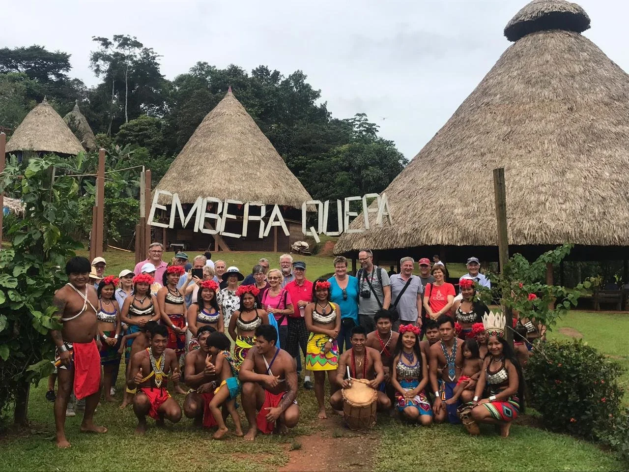 Tourists and indigenous people gathering in front of traditional thatched huts, with a sign that reads "Embera Gua" in the background, at an ethnic village or cultural site.