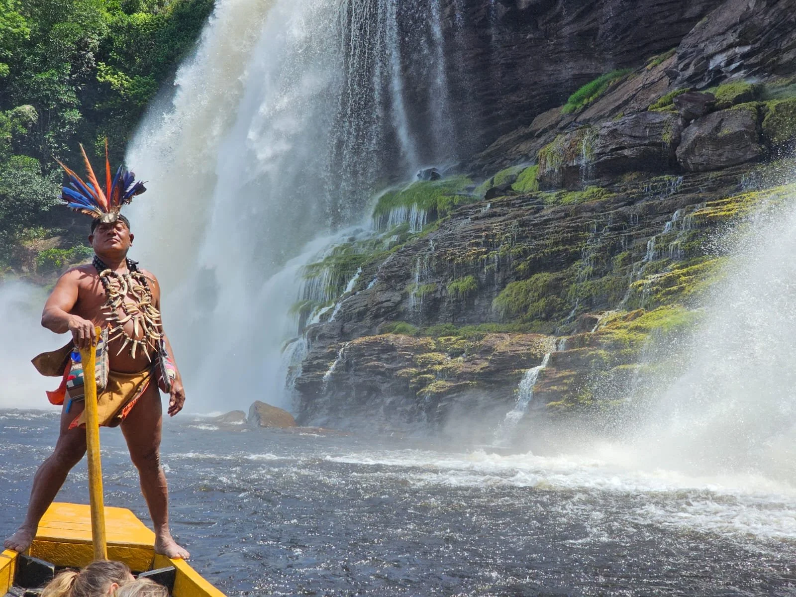 A Maori man standing on a boat in front of a waterfall, dressed in traditional Maori attire with a feathered headpiece and necklaces, holding a staff.