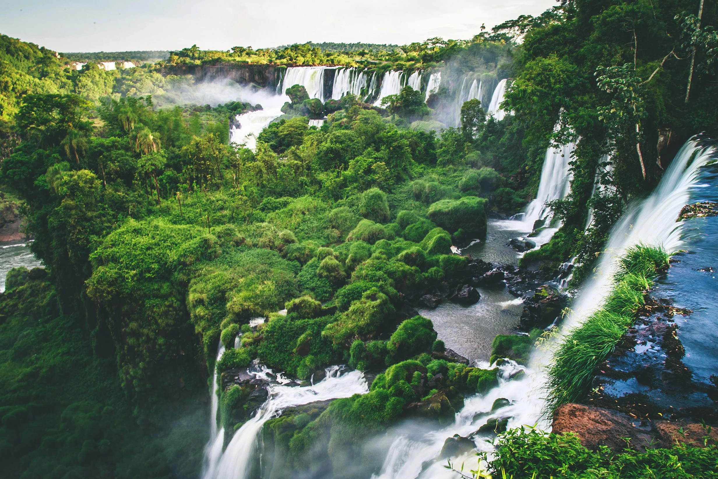 A lush green landscape with cascading waterfalls flowing into a river, surrounded by dense trees and vegetation under a partly cloudy sky.