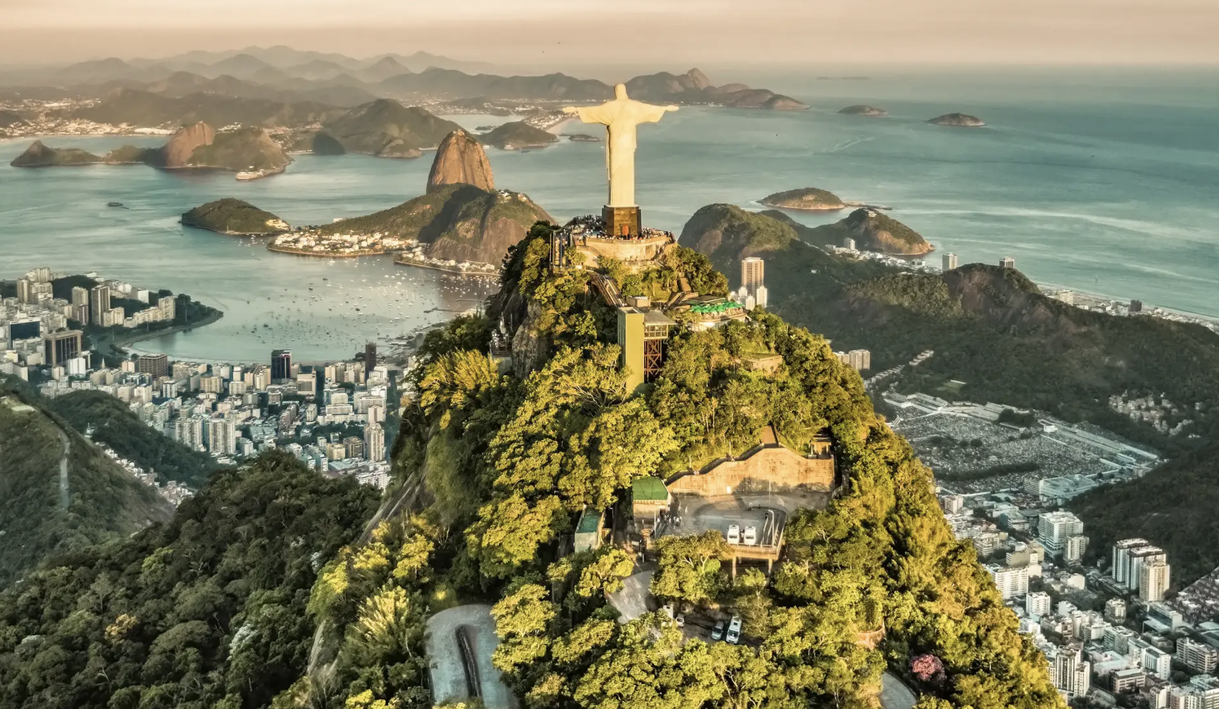 Aerial view of the Christ the Redeemer statue atop Corcovado Mountain in Rio de Janeiro, Brazil, with surrounding cityscape, coastline, and ocean in the distance during sunset.