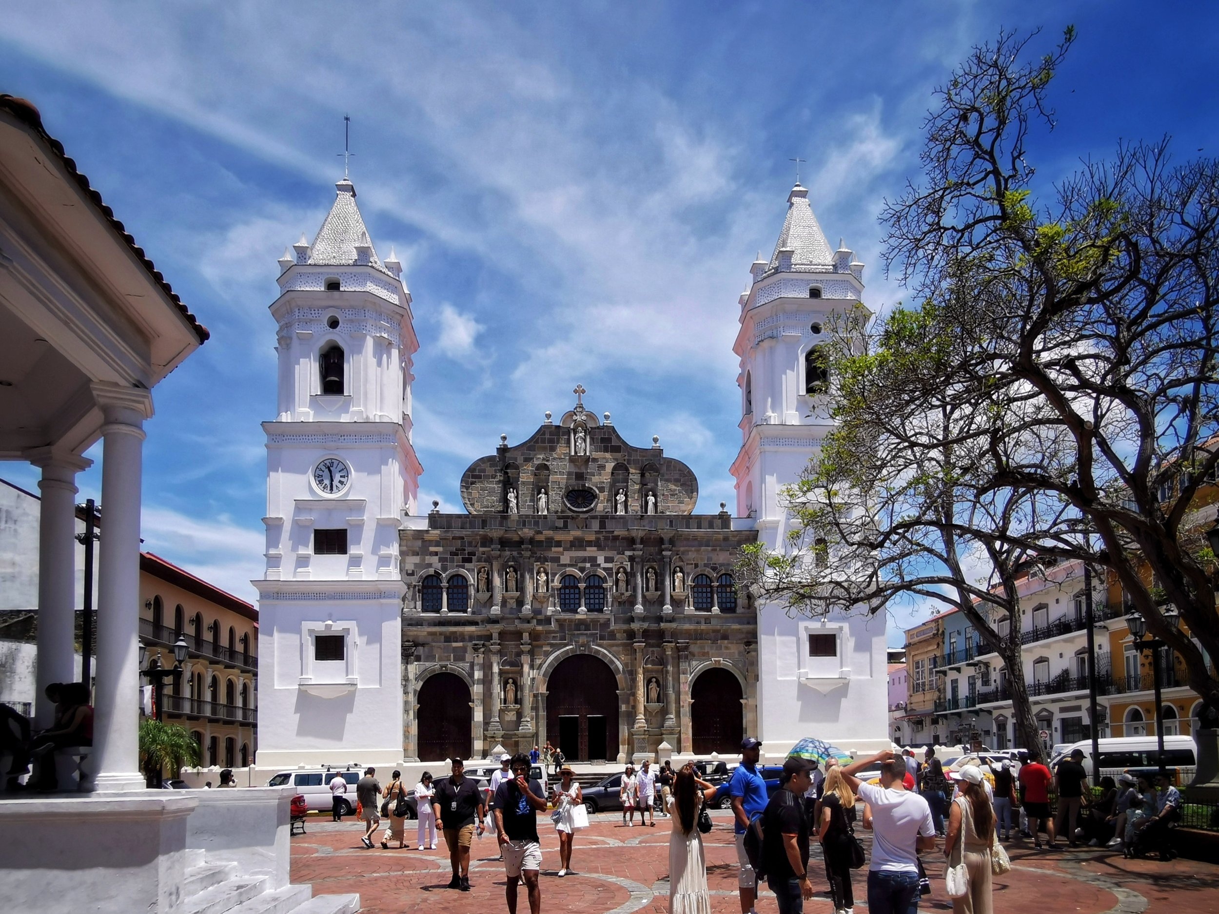 The Basilica of Our Lady of Guadalupe in Mexico City with people walking and gathering in the plaza ahead, blue sky with scattered clouds, a tree with sparse leaves on the right, and surrounding buildings.