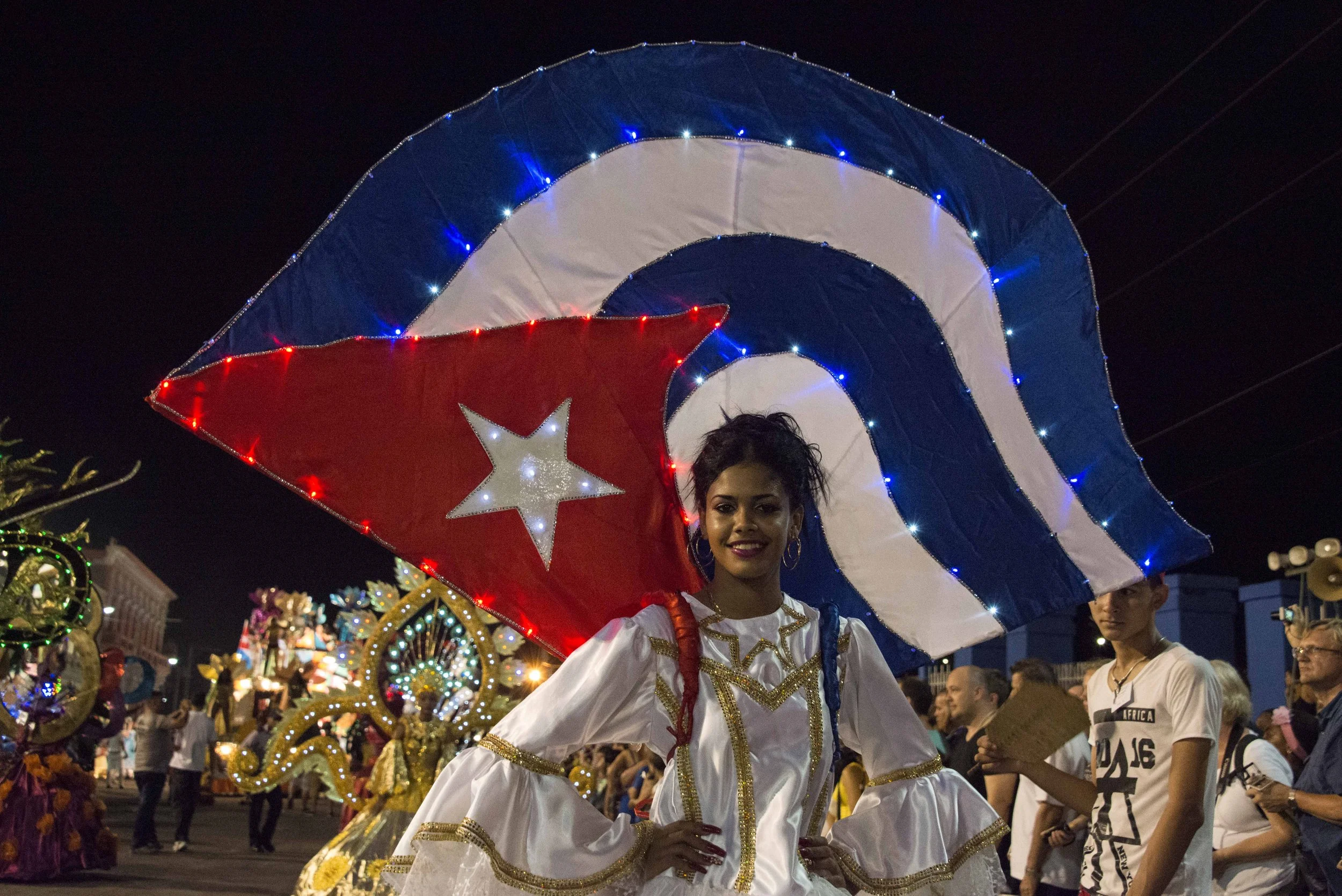 A young woman in a white dress with gold accents is smiling at a night-time parade. She stands under an illuminated flag of Cuba with red, white, and blue colors, and a white star. She is surrounded by other parade participants and spectators.