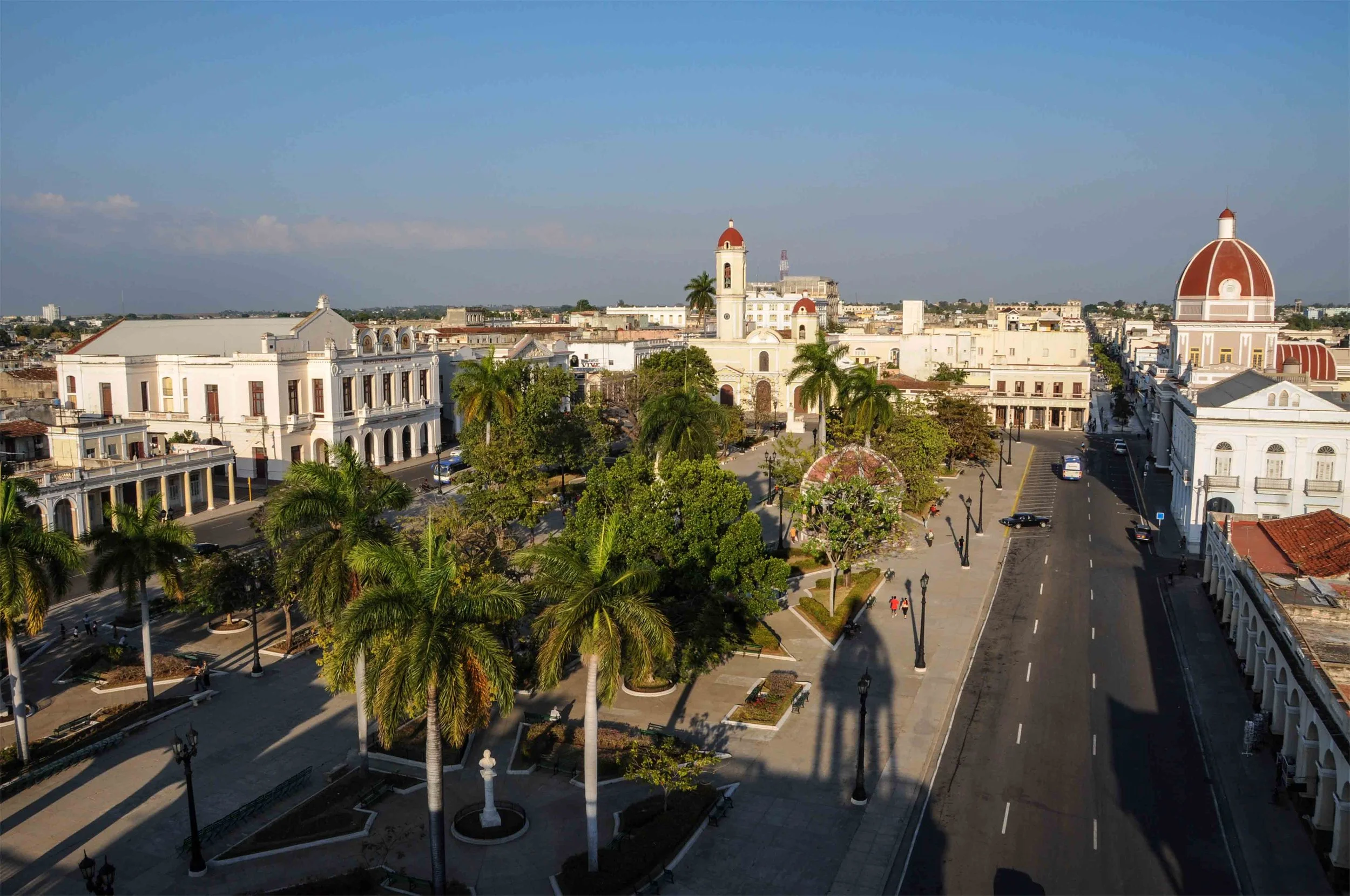 Aerial view of a city square with historic white buildings, palm trees, a park, and a few pedestrians on the sidewalks, under a clear blue sky. Cuba.