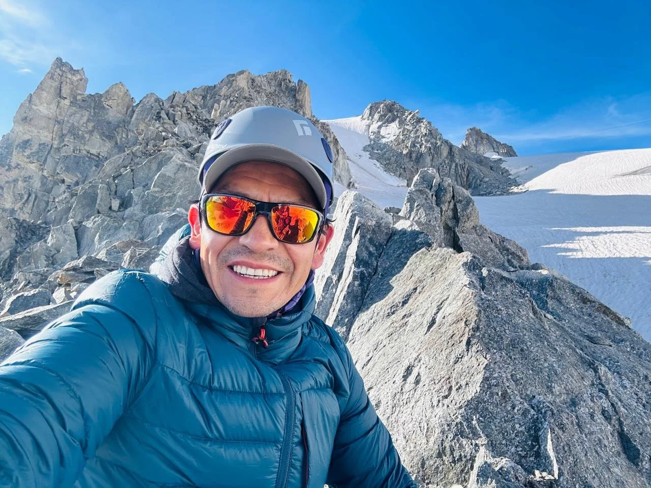 A man in outdoor gear, including a helmet and sunglasses, taking a selfie on a snowy mountain with rocky terrain and snow-covered peaks under a blue sky in Ecuador.
