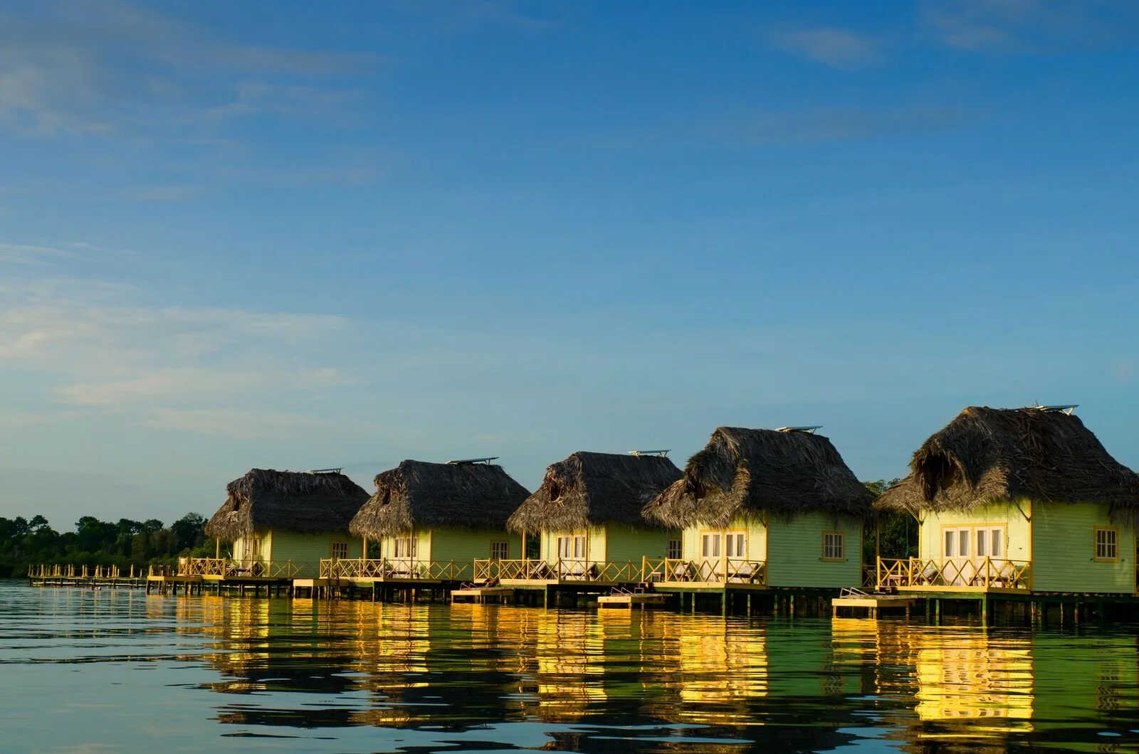 Overwater cottages with thatched roofs and wooden railings along a calm body of water, with trees in the background and a blue sky with some clouds.