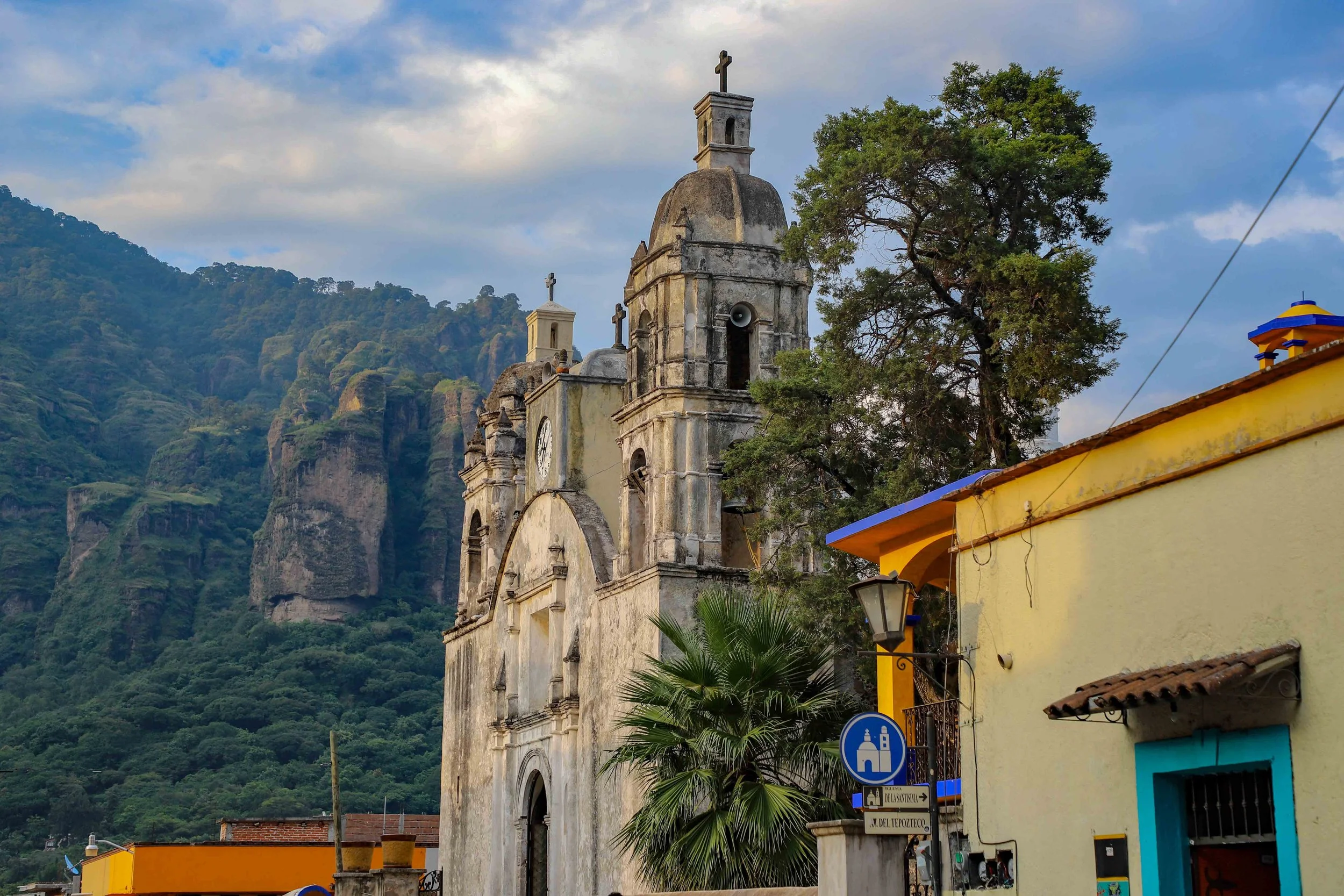 An old church with tall bell towers and a cross at the top, set against a backdrop of green mountains. A palm tree and colorful buildings are in the foreground, with a cloudy sky overhead.