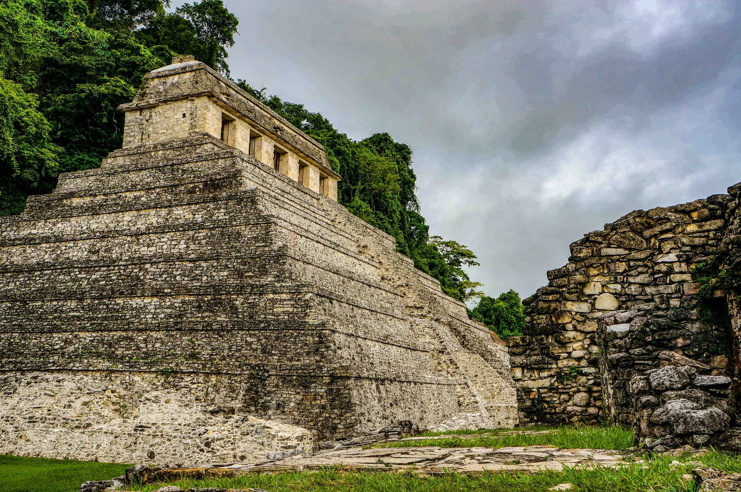 Ancient stone pyramid with a rectangular structure and multiple terraces, surrounded by greenery and cloudy skies.