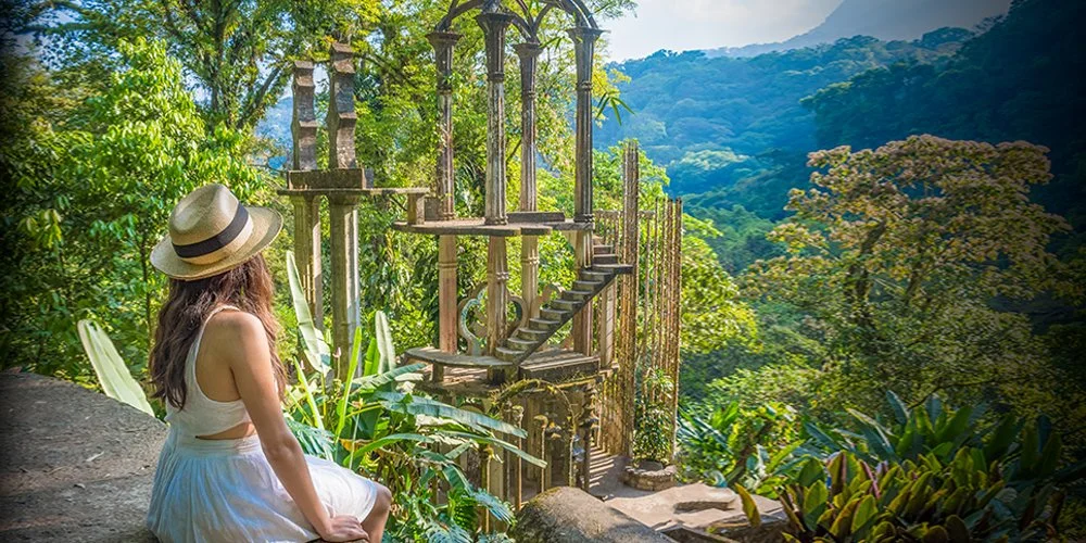 A woman wearing a dress and a straw hat is sitting on a rock, looking at a floating staircase structure in a lush green jungle with mountains in the background.