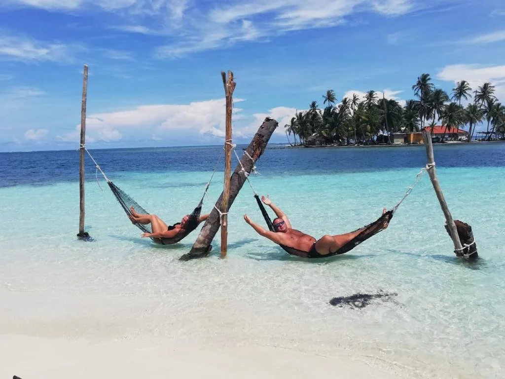Two people relaxing in hammocks tied to wooden poles on a tropical beach with clear turquoise water, palm trees, and a partly cloudy sky in the background.