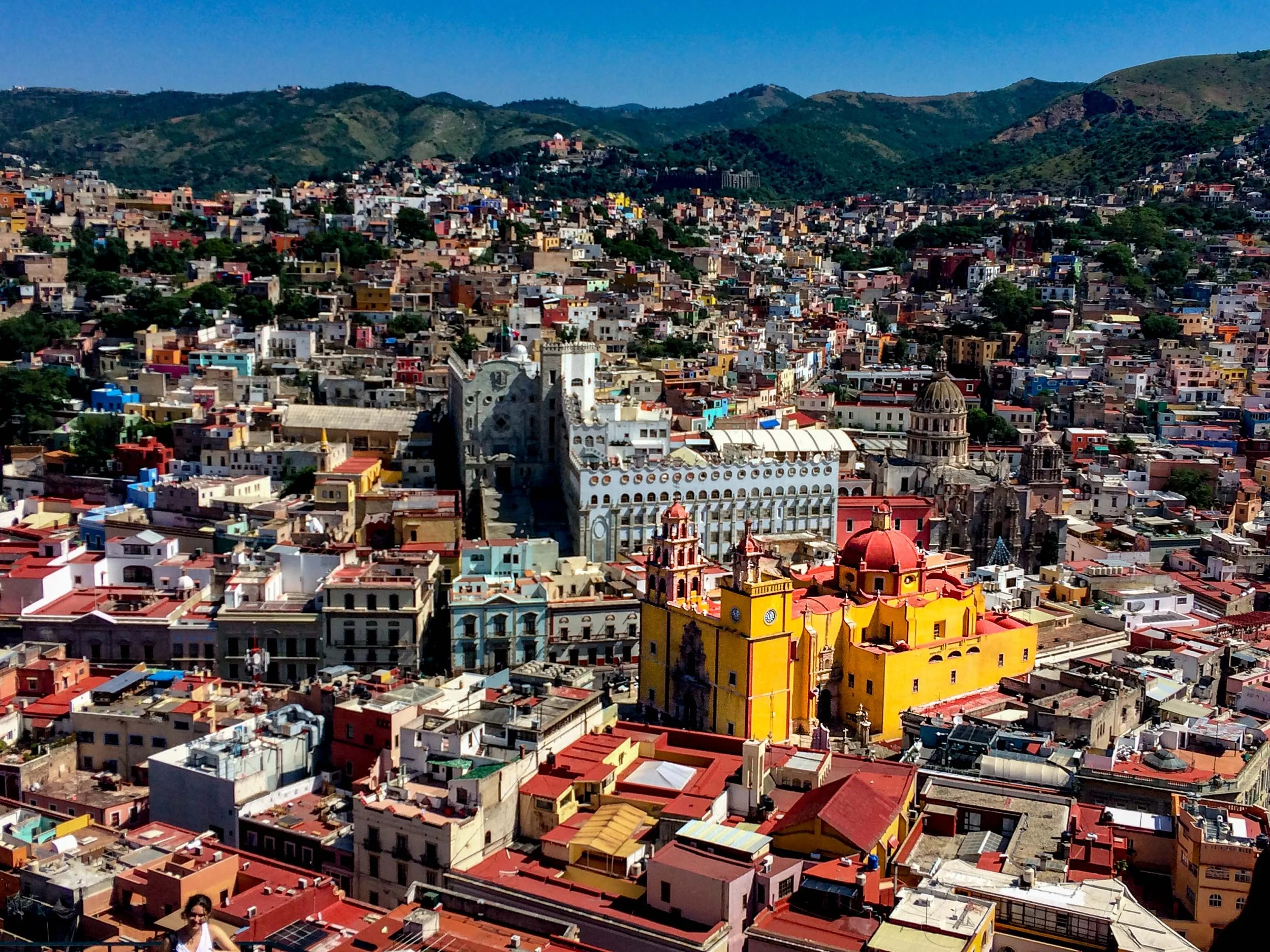 A vibrant cityscape of Mexico City with colorful buildings, historic architecture, and green hills in the background under a clear blue sky.