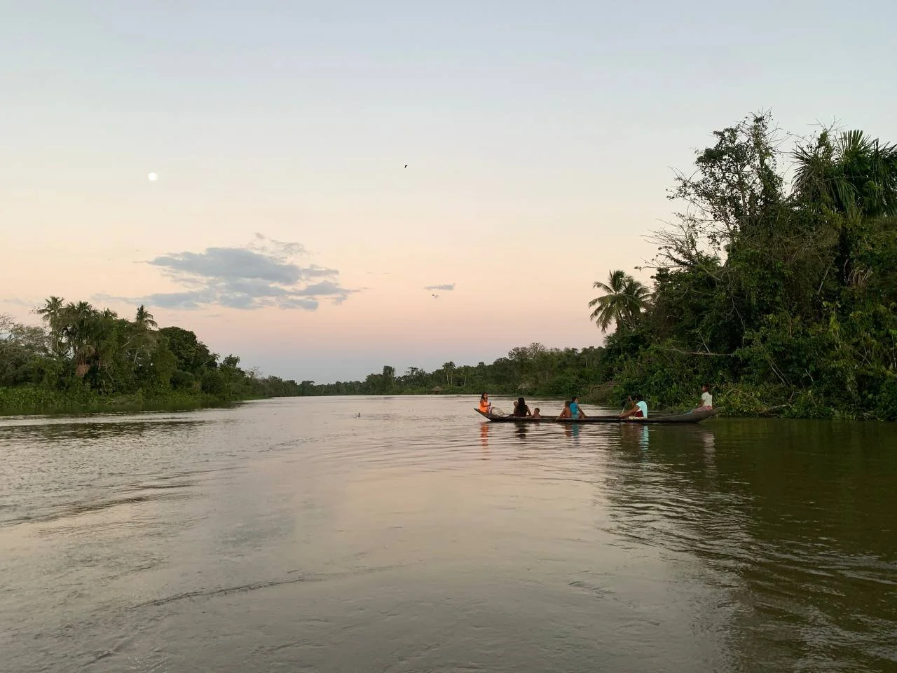 A group of people are riding a canoe on a calm river during dusk, surrounded by trees and vegetation.