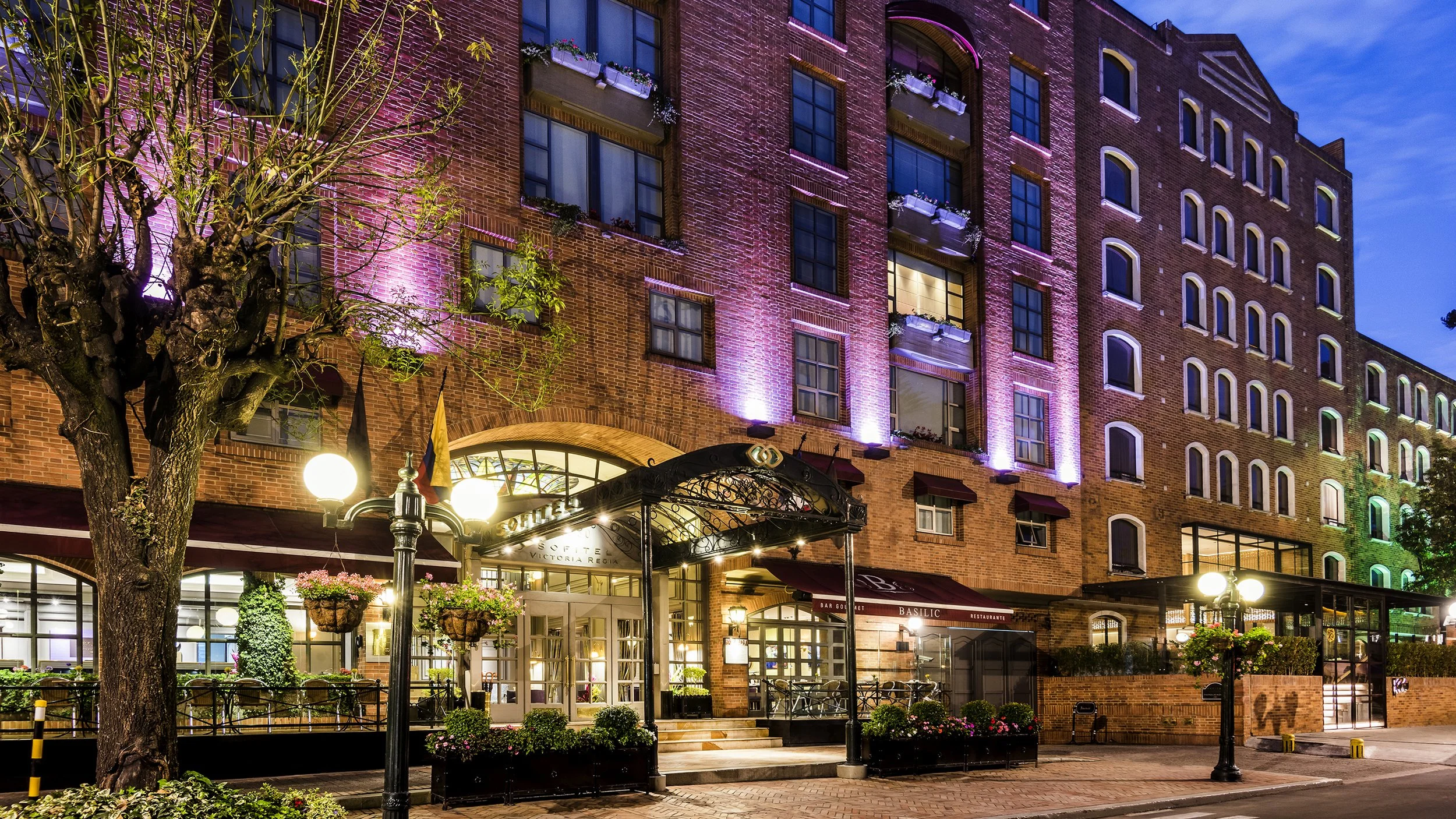 A brightly lit hotel entrance at dusk, with colorful exterior lighting on a brick building, trees, hanging flower baskets, and street lamps.
