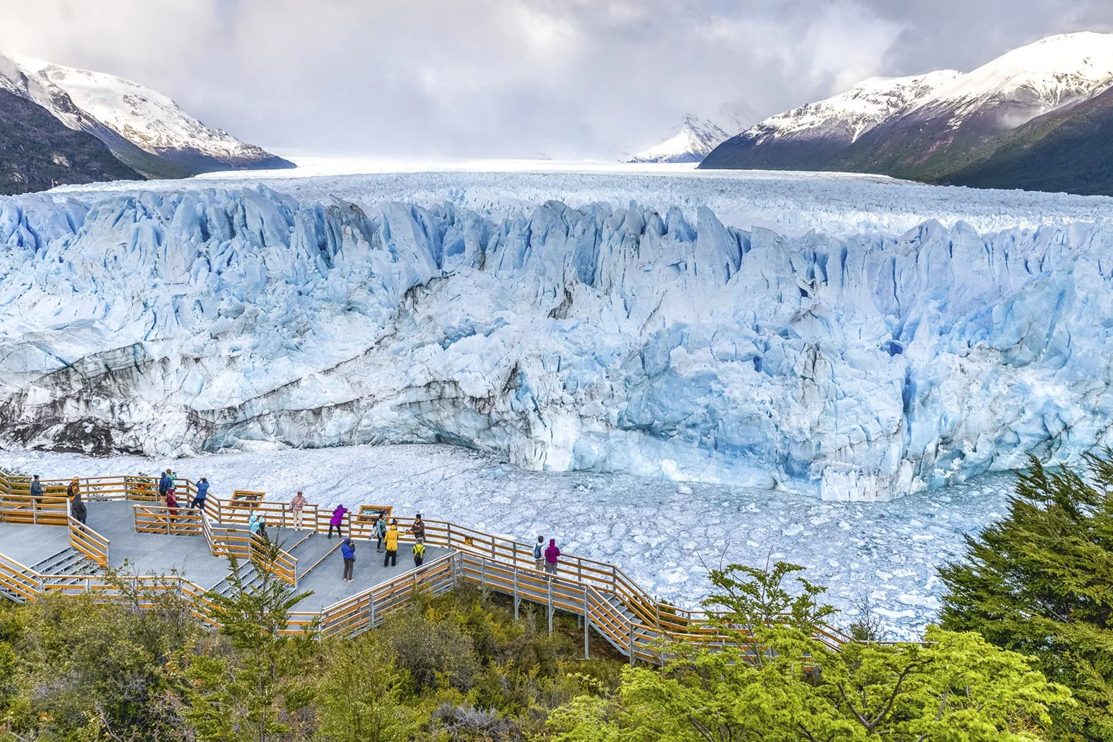 Tourists observing a glacier from a viewing platform surrounded by greenery with snow-capped mountains in the background.