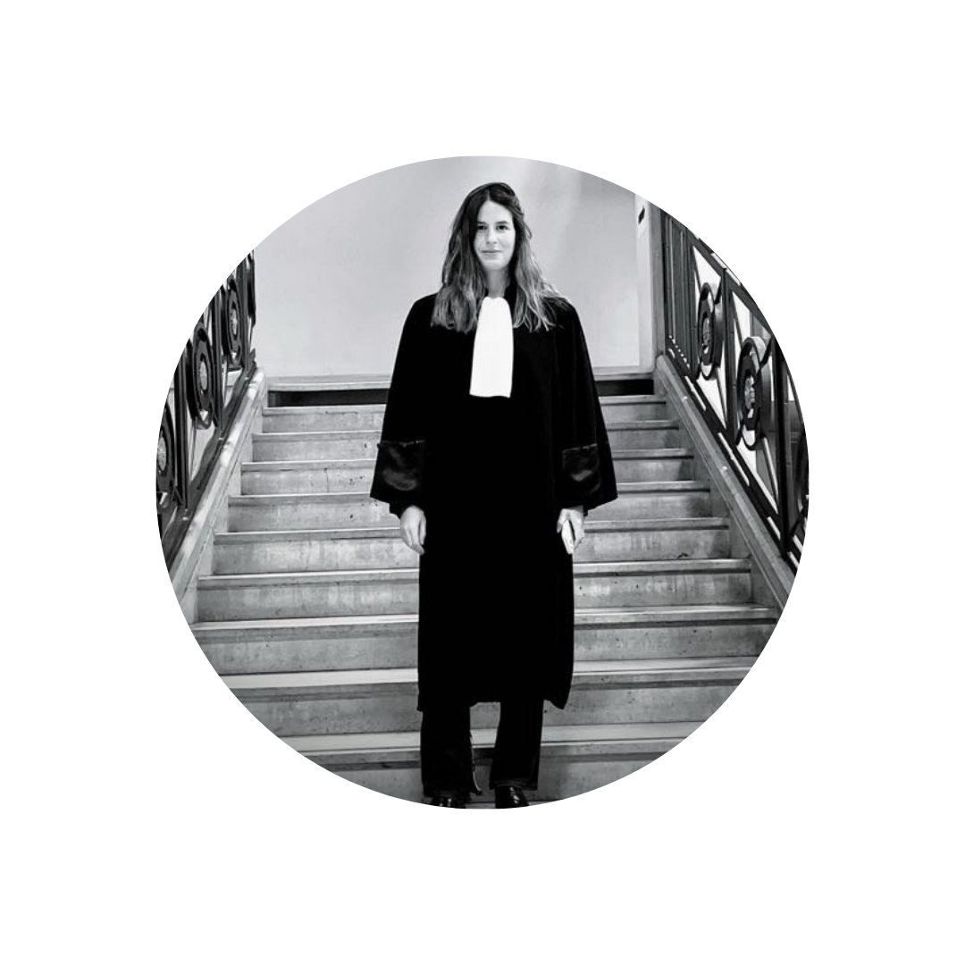 Une femme en robe de juge, posant en haut d'un escalier en bois avec des rampes en métal, dans une salle en intérieur.