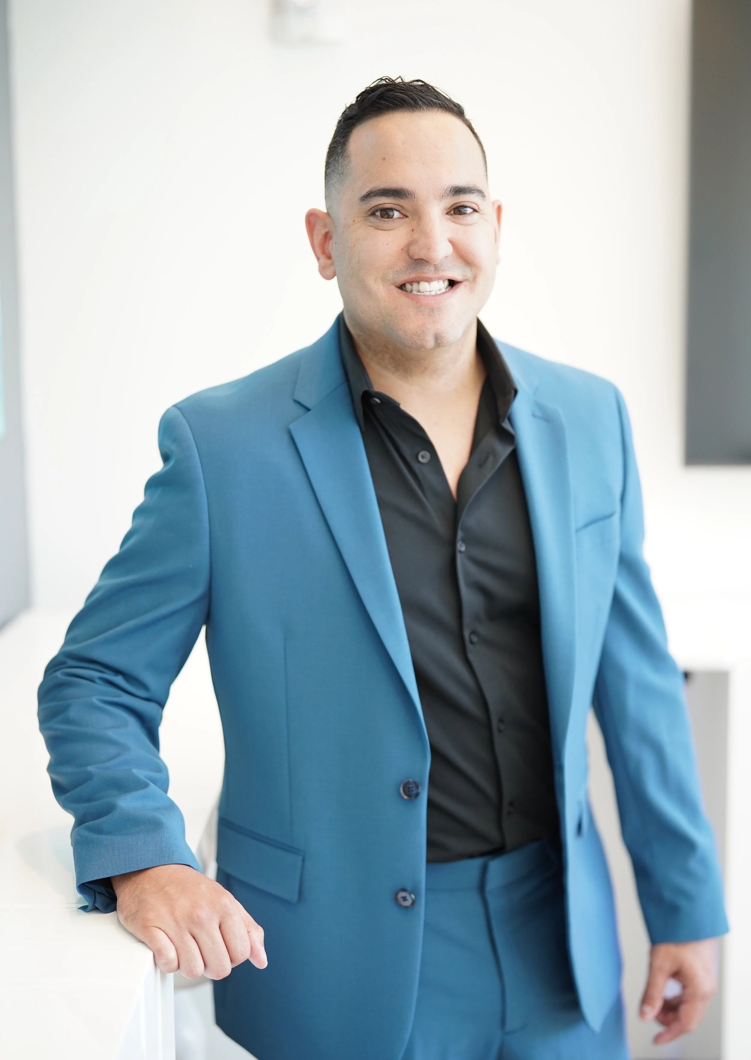 A man in a blue suit and black shirt smiling and standing indoors.