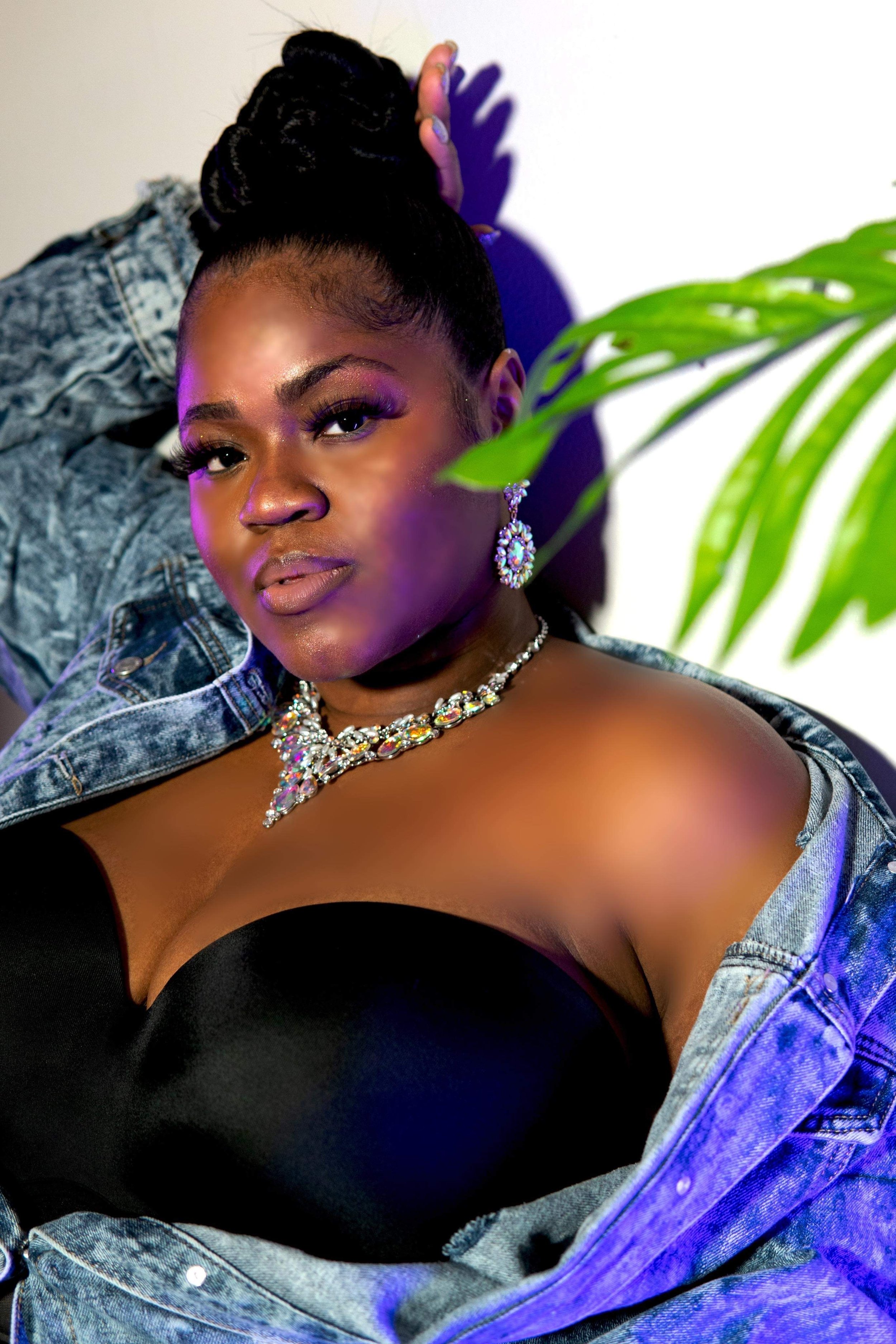 Woman in a denim jacket with a black strapless top, wearing statement jewelry, poses next to a green plant.