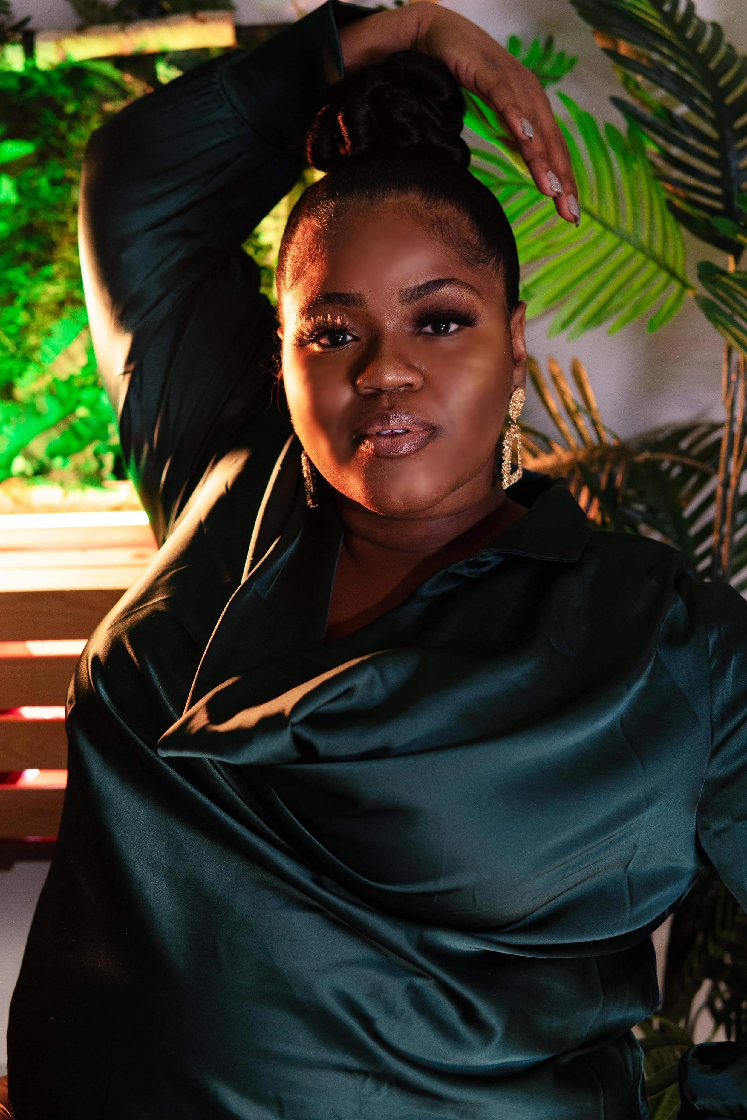 Person posing with arm raised, wearing a dark green satin outfit, with green plants in the background.