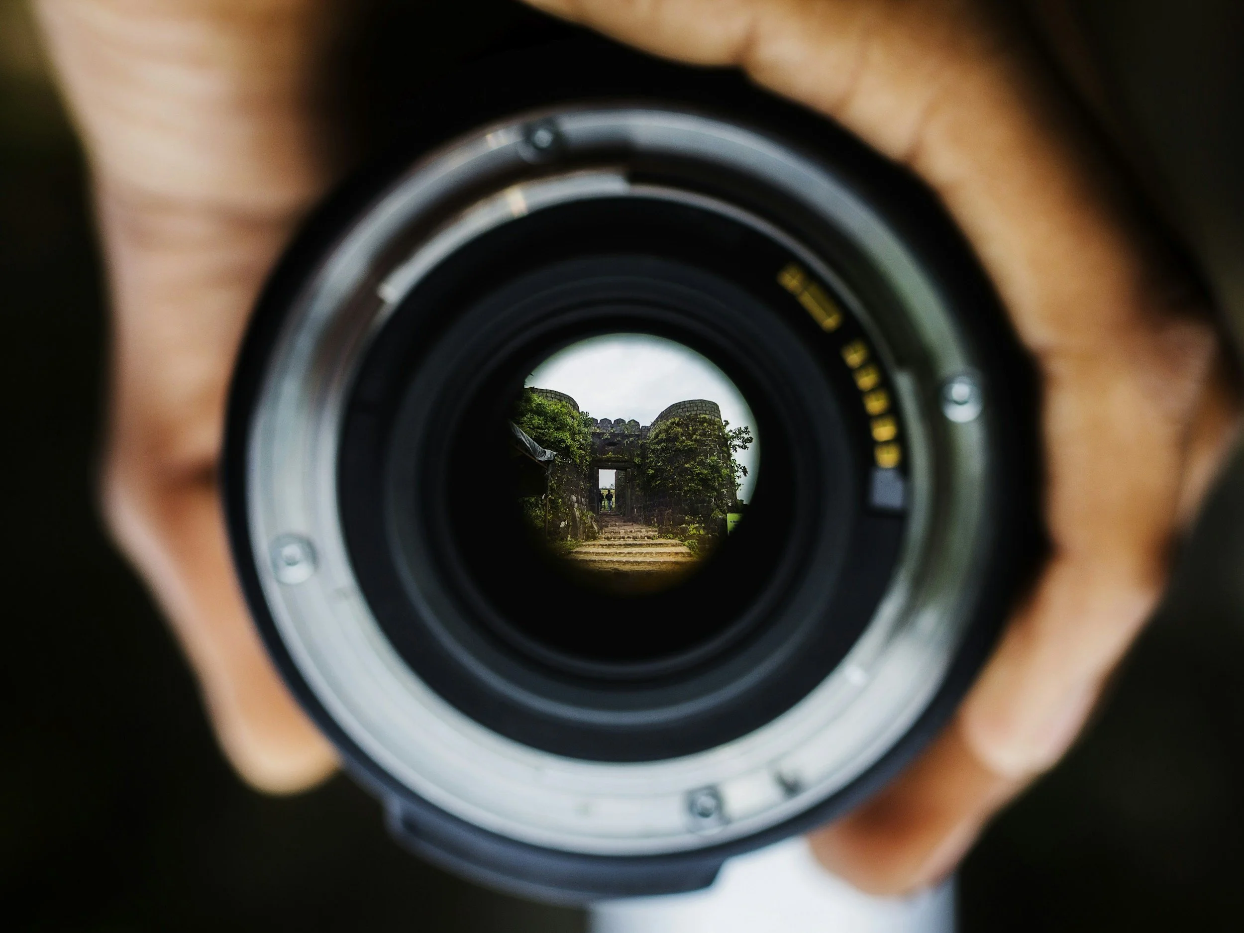 A person holding a camera lens, showing a small, focused view of a stone structure and greenery through the lens.