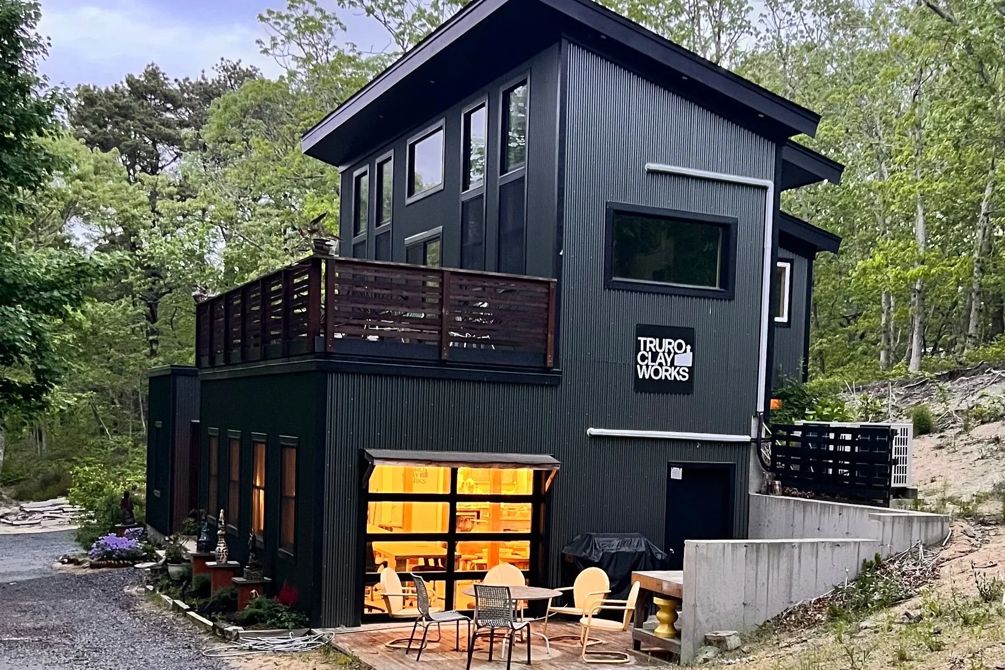 Modern black house with multiple levels and large windows surrounded by trees, with outdoor seating area and a sign that reads "Truro Clay Works".