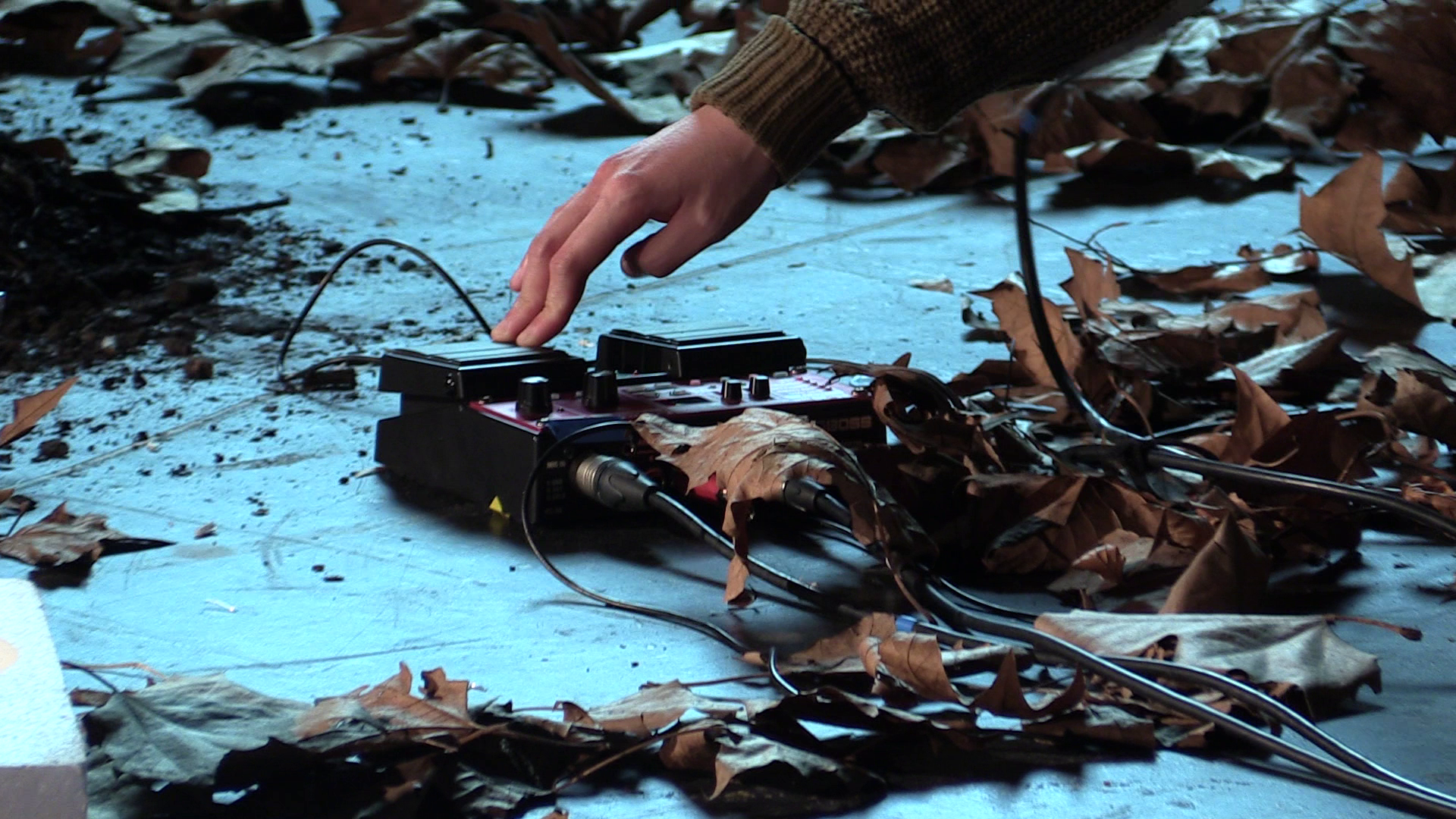 A person's hand adjusts electronic equipment on the floor surrounded by dried leaves.