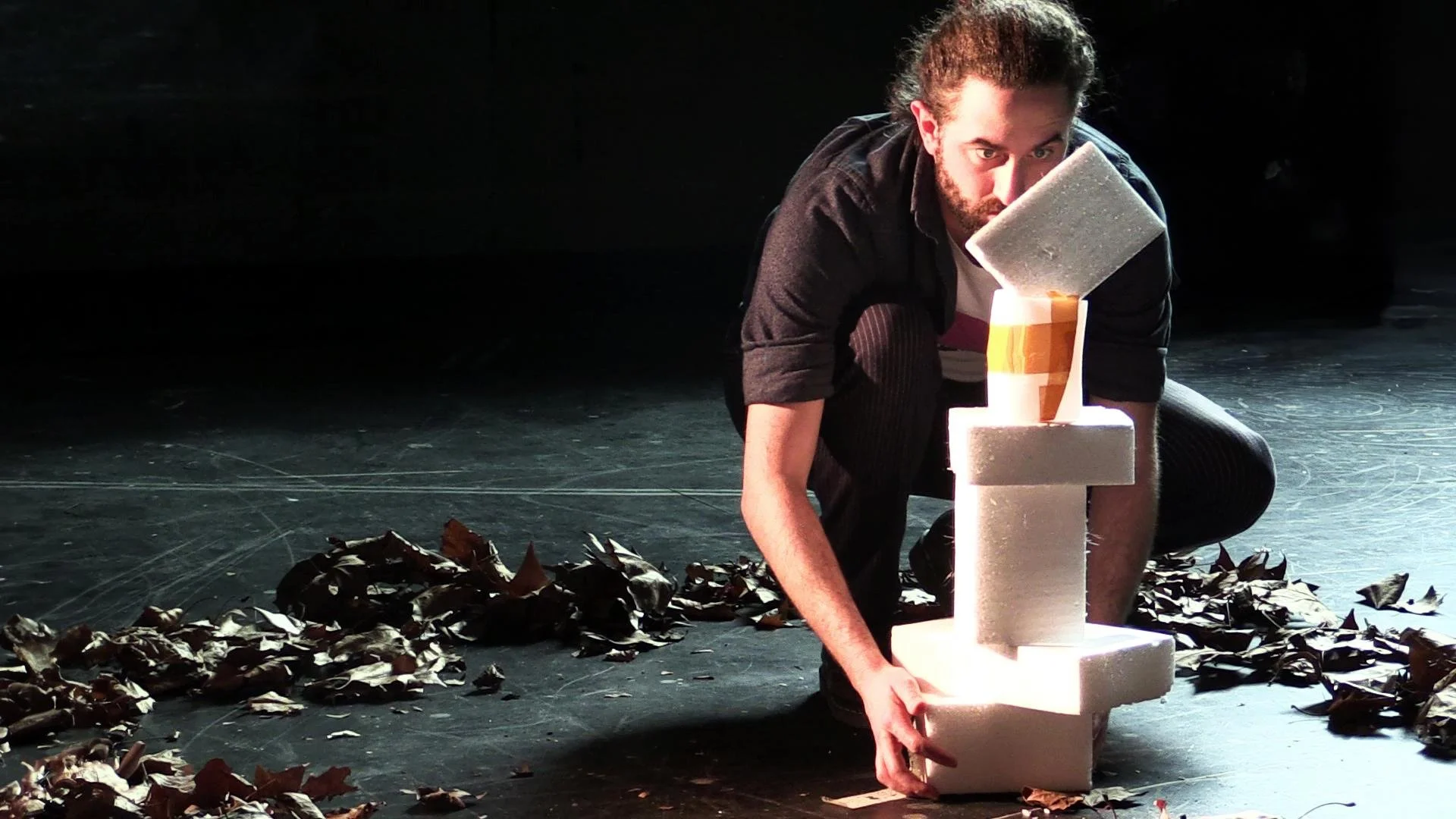 Man with curly hair and beard crouching down, assembling a tower of white foam blocks, with a paper cup on top, on a dark stage with brown leaves scattered around.
