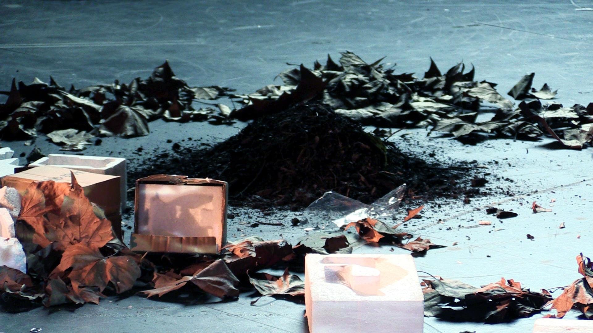 A pile of soil and ashes surrounded by scattered dried leaves and boxes on a flat surface.