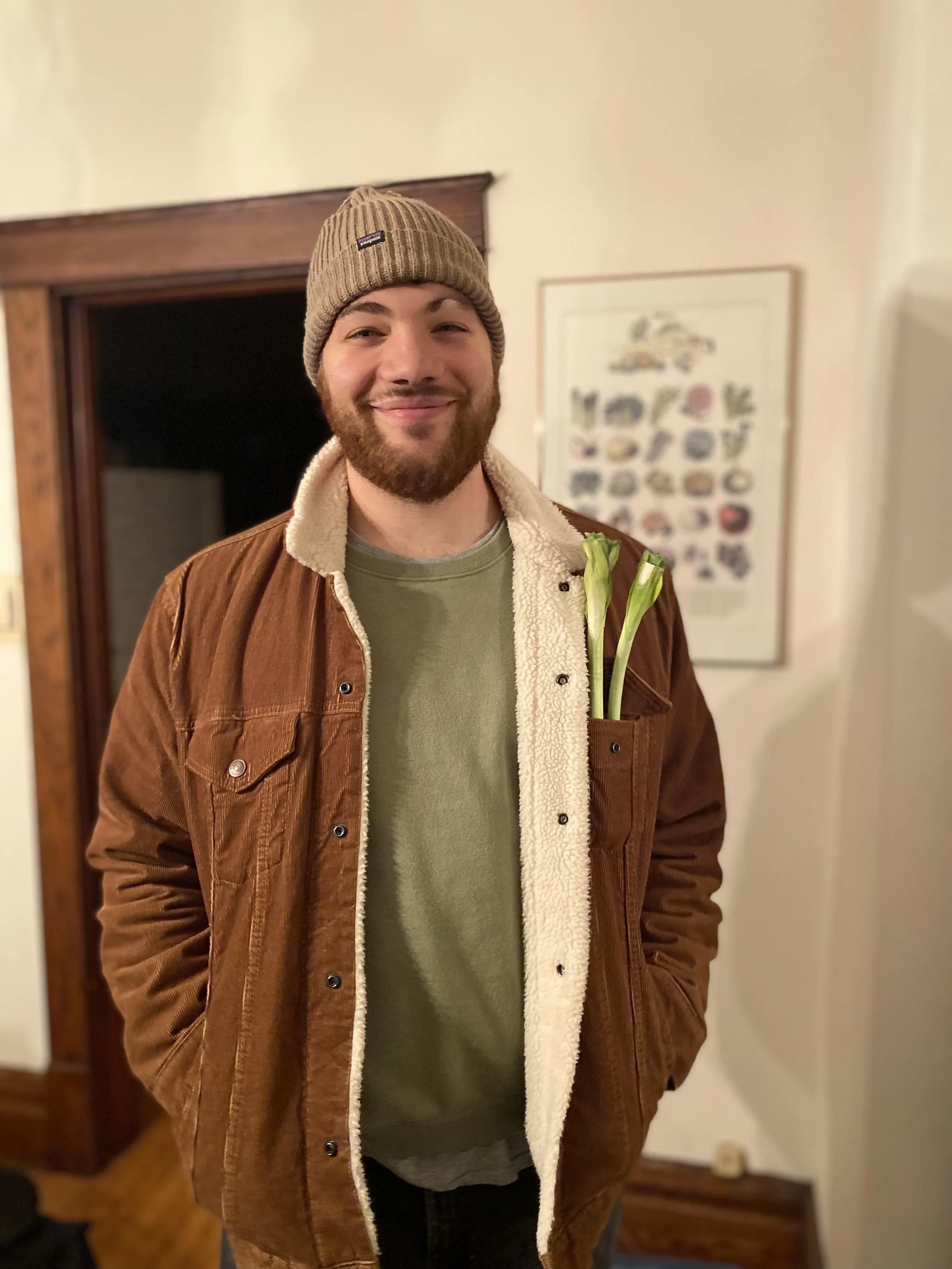 A smiling man with a beard wearing a tan beanie, khaki shirt, and brown jacket with a cream lining, standing indoors with two green calla lilies in his shirt pocket.