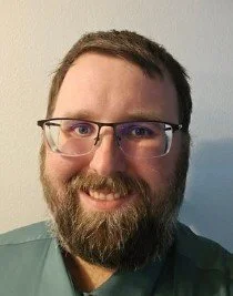 Portrait of a man with glasses, a beard, and short dark hair, smiling in front of a light-colored wall.