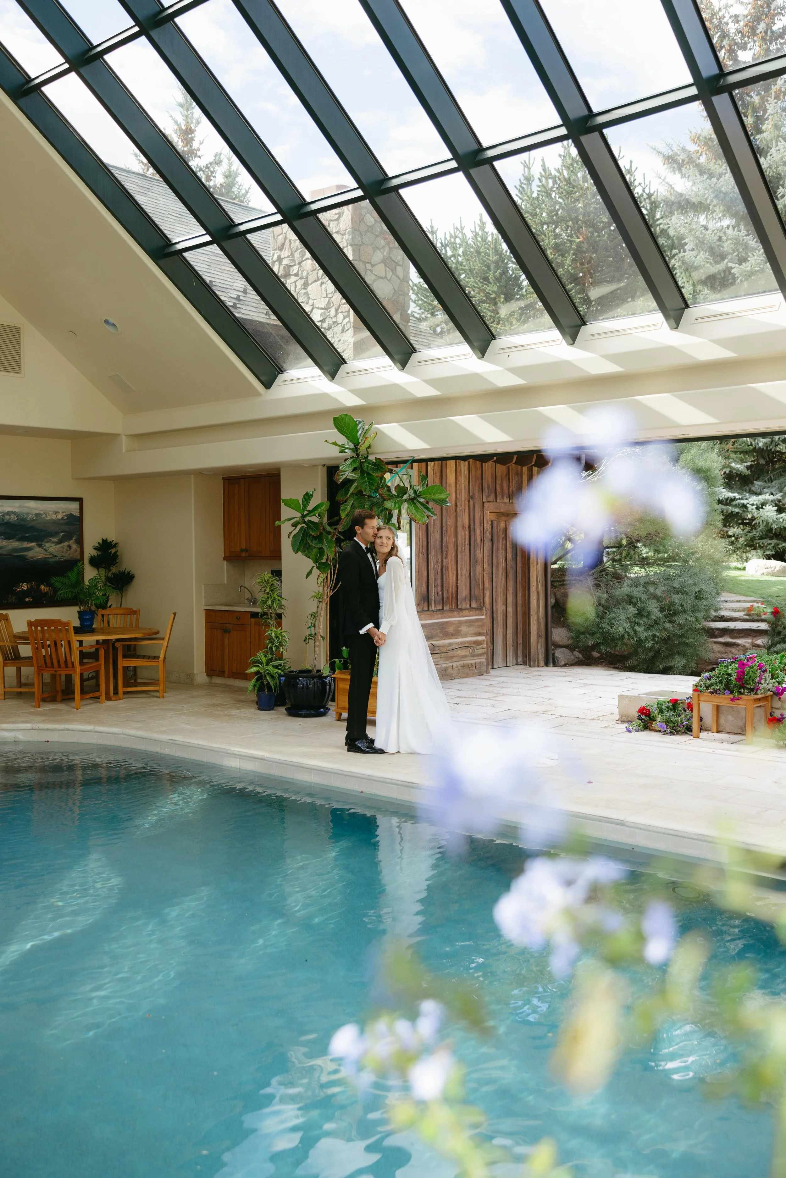 A bride and groom standing hand in hand indoors next to a swimming pool, with sunlight coming through a large glass ceiling, and plants and flowers in the background.
