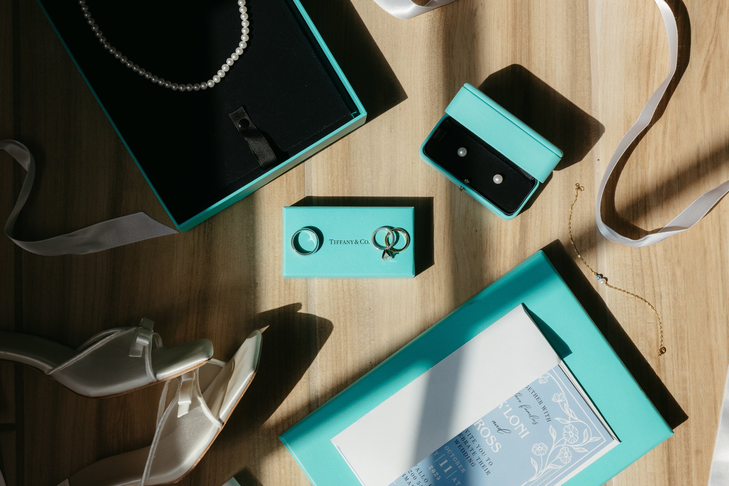 Jewelry and accessories on a wooden surface, including rings, earrings in Tiffany blue boxes, a pearl necklace, a bracelet, and a pair of white satin shoes.